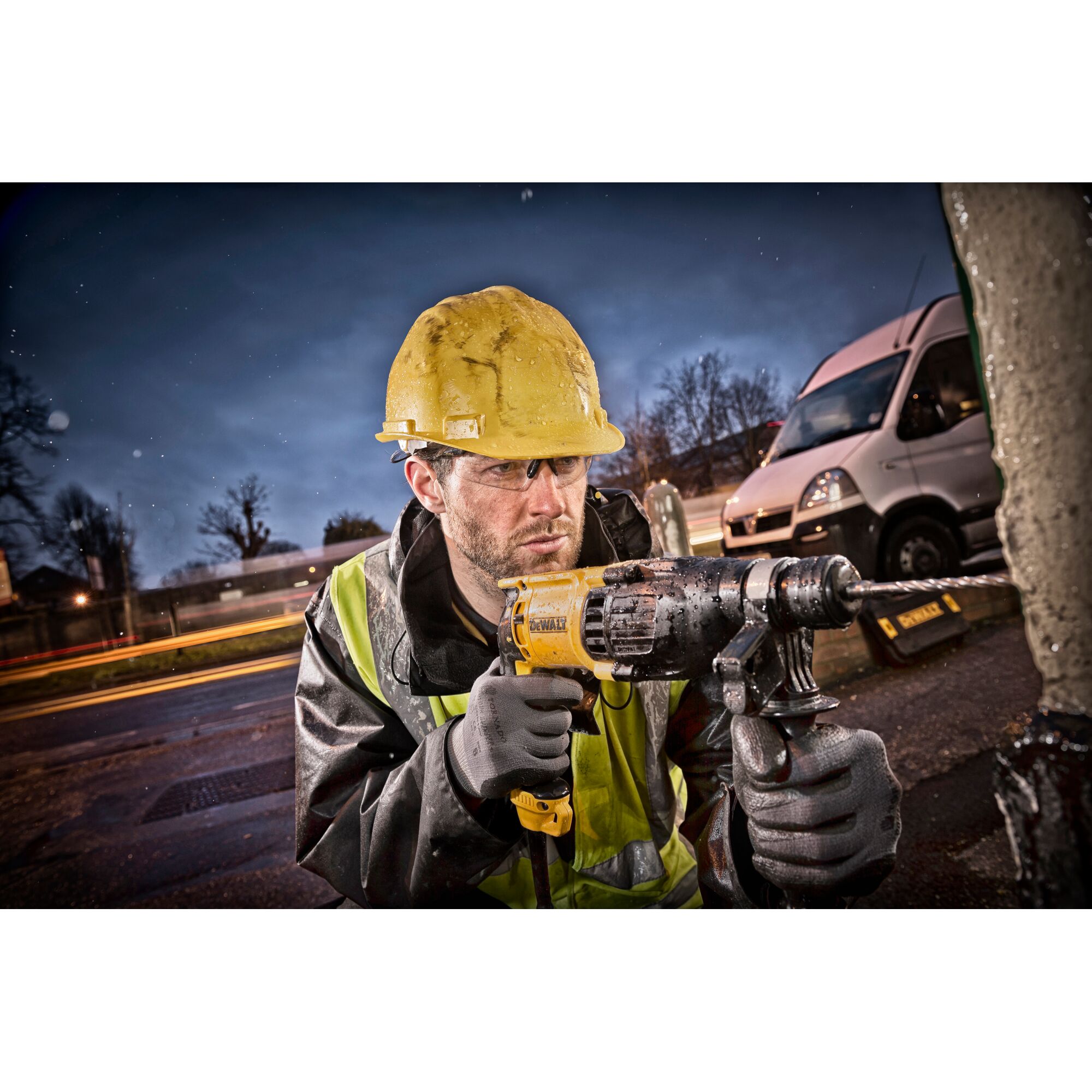 A person wearing a yellow hard hat and reflective safety vest uses a DEWALT electric drill outdoors, with a van and blurred lights visible in the background. The person's face is obscured.