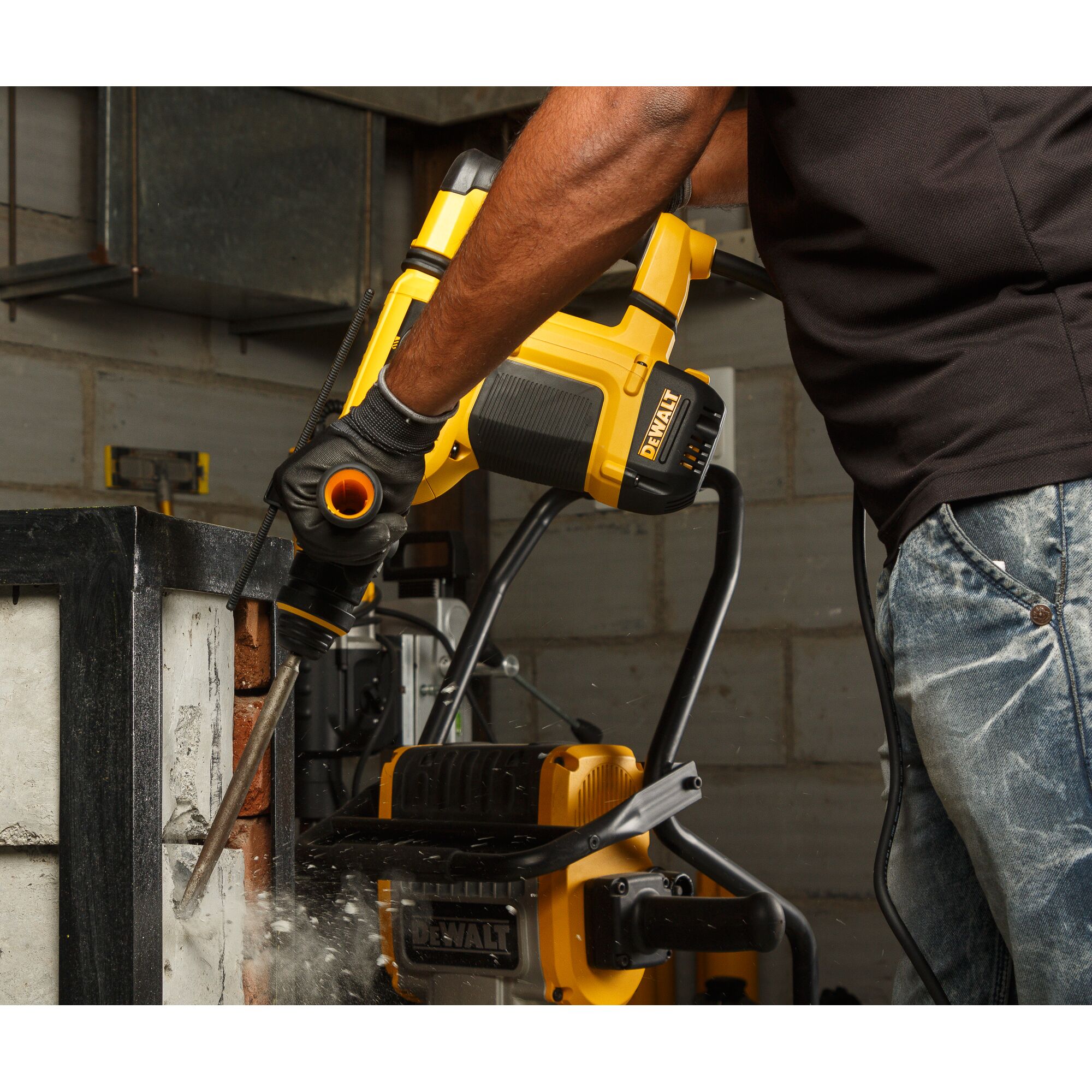 A person using a DEWALT rotary hammer drill to chisel concrete in an industrial setting. Dust and debris are visible around the tool during operation.