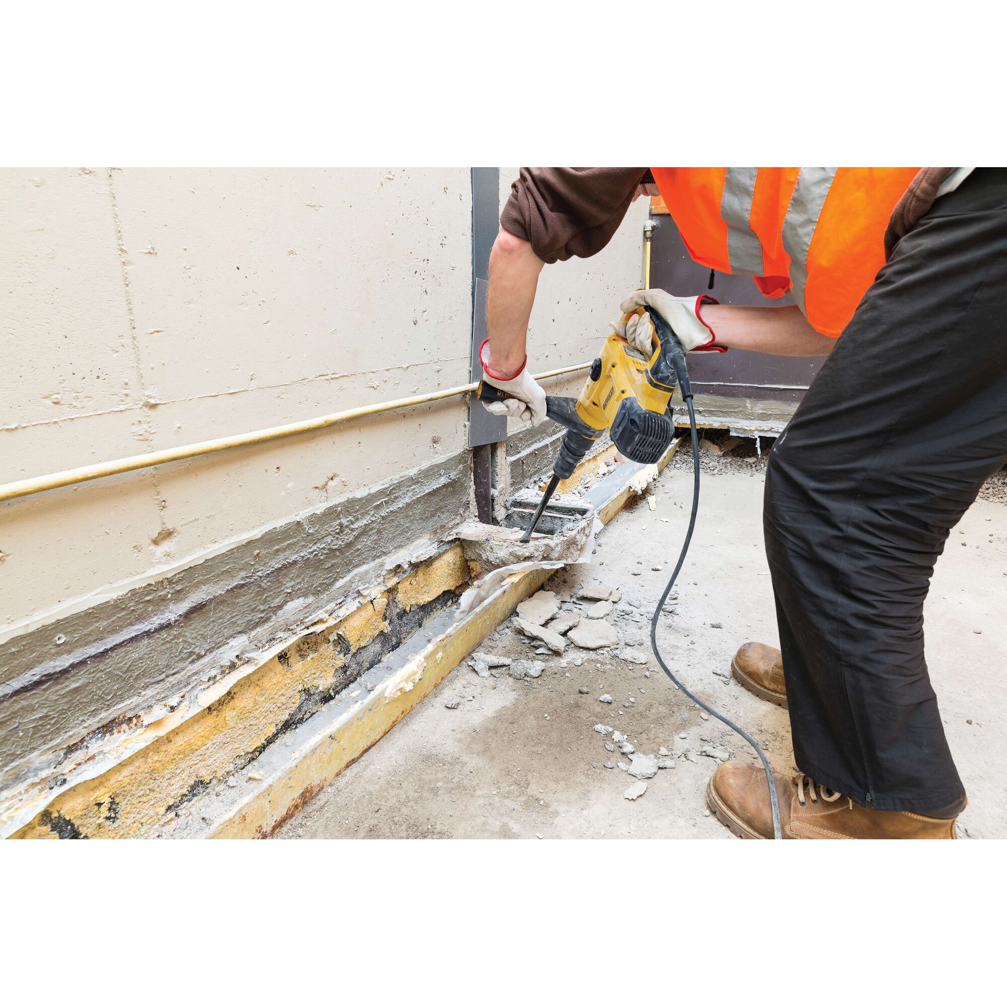 A person wearing safety gear uses a yellow DEWALT demolition hammer to break concrete along the base of a wall outdoors. Pieces of broken concrete and dust are visible on the ground.