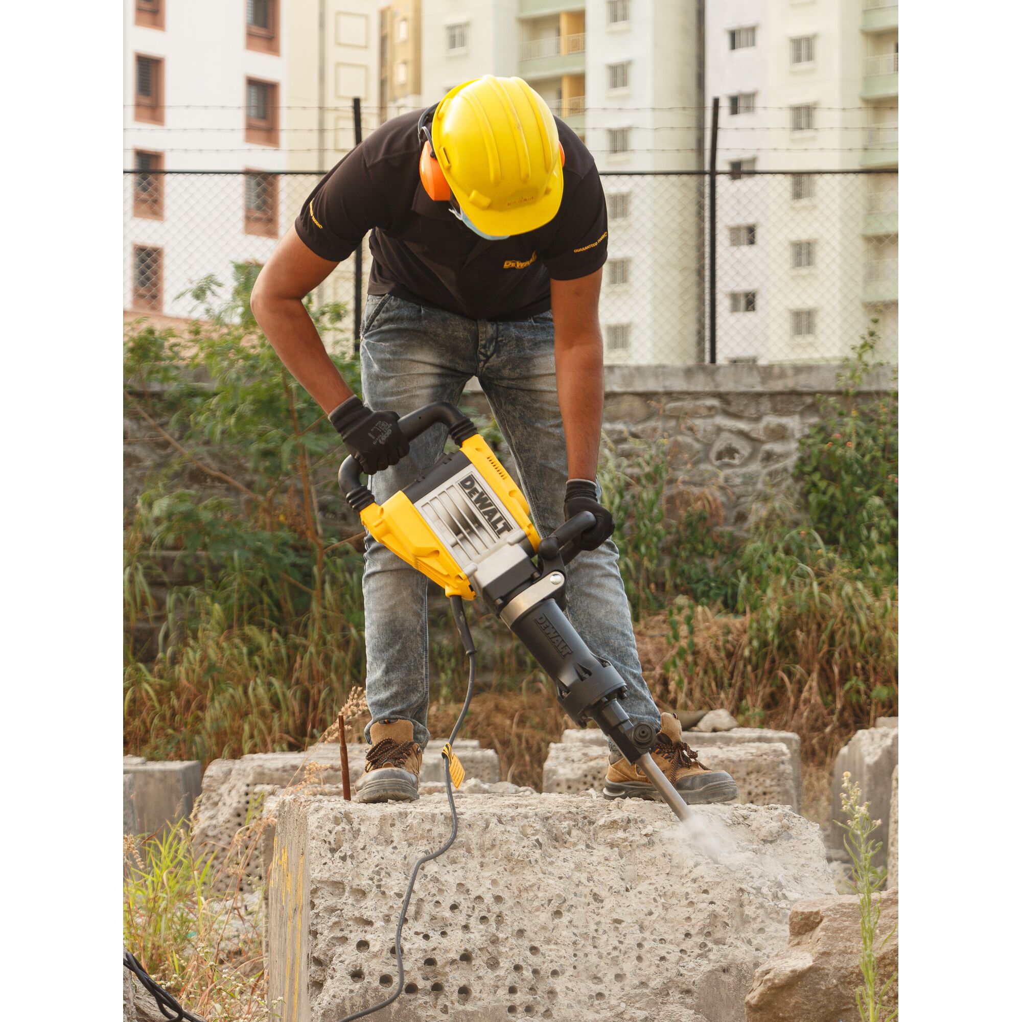A person wearing safety gear uses a DEWALT D25961K demolition hammer to break a large concrete block at a construction site outdoors.