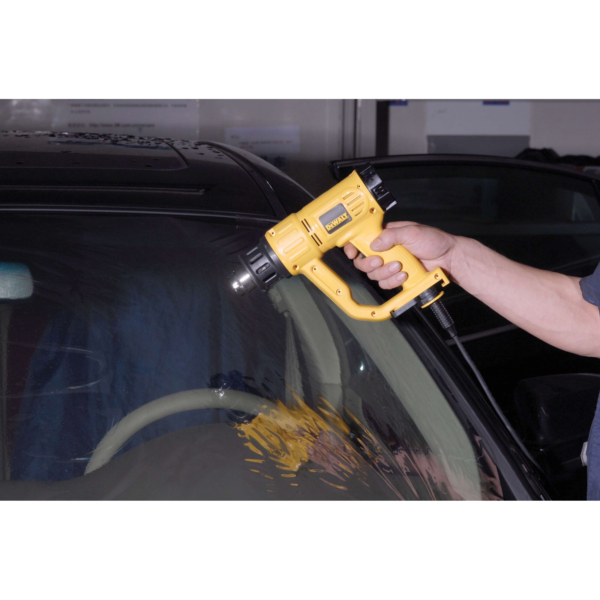 A person using a yellow DEWALT heat gun (model D26411) to apply or remove material on a car windshield.