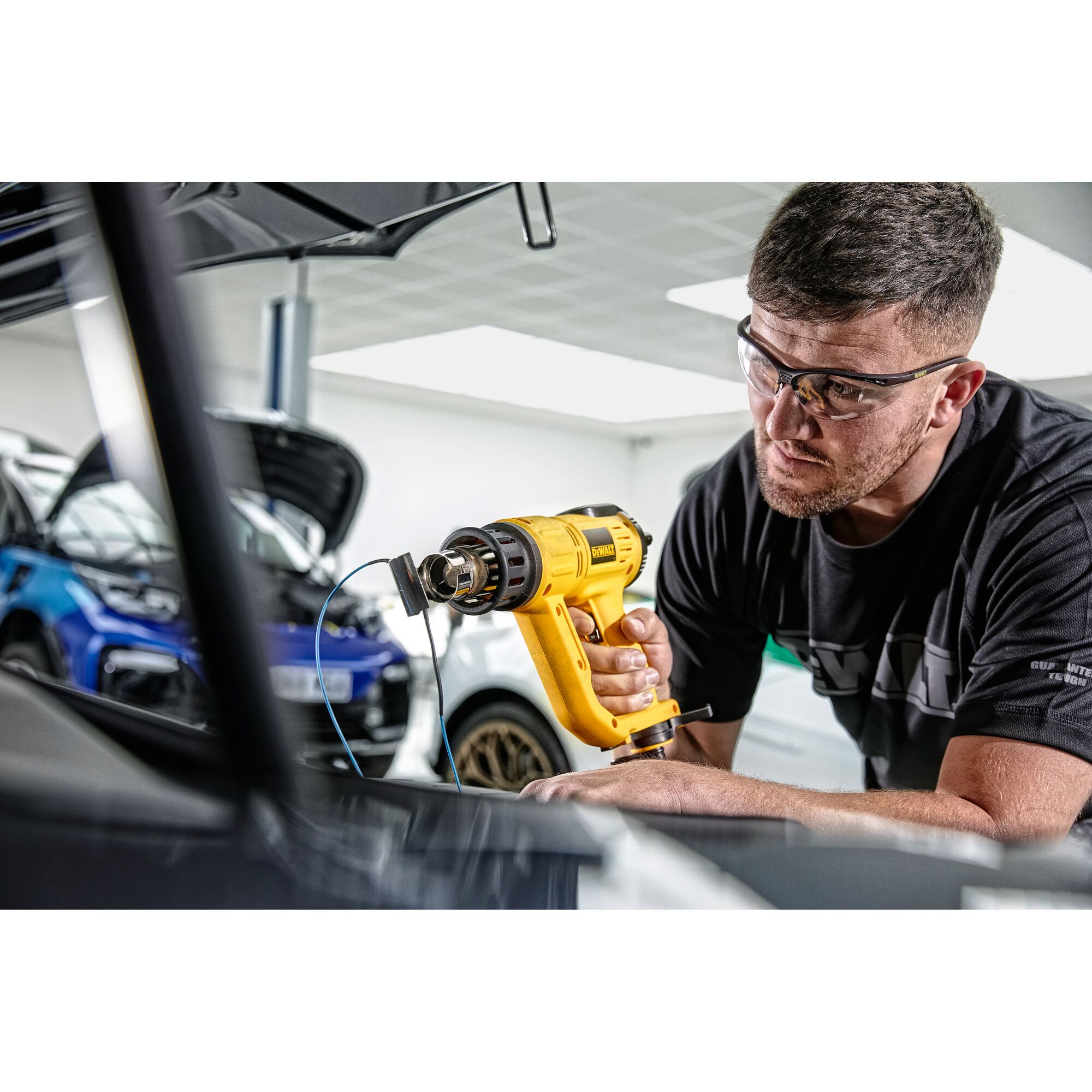 A person uses a DEWALT D26414 yellow heat gun in a garage or workshop, with cars visible in the background. The person’s face is blurred for privacy.