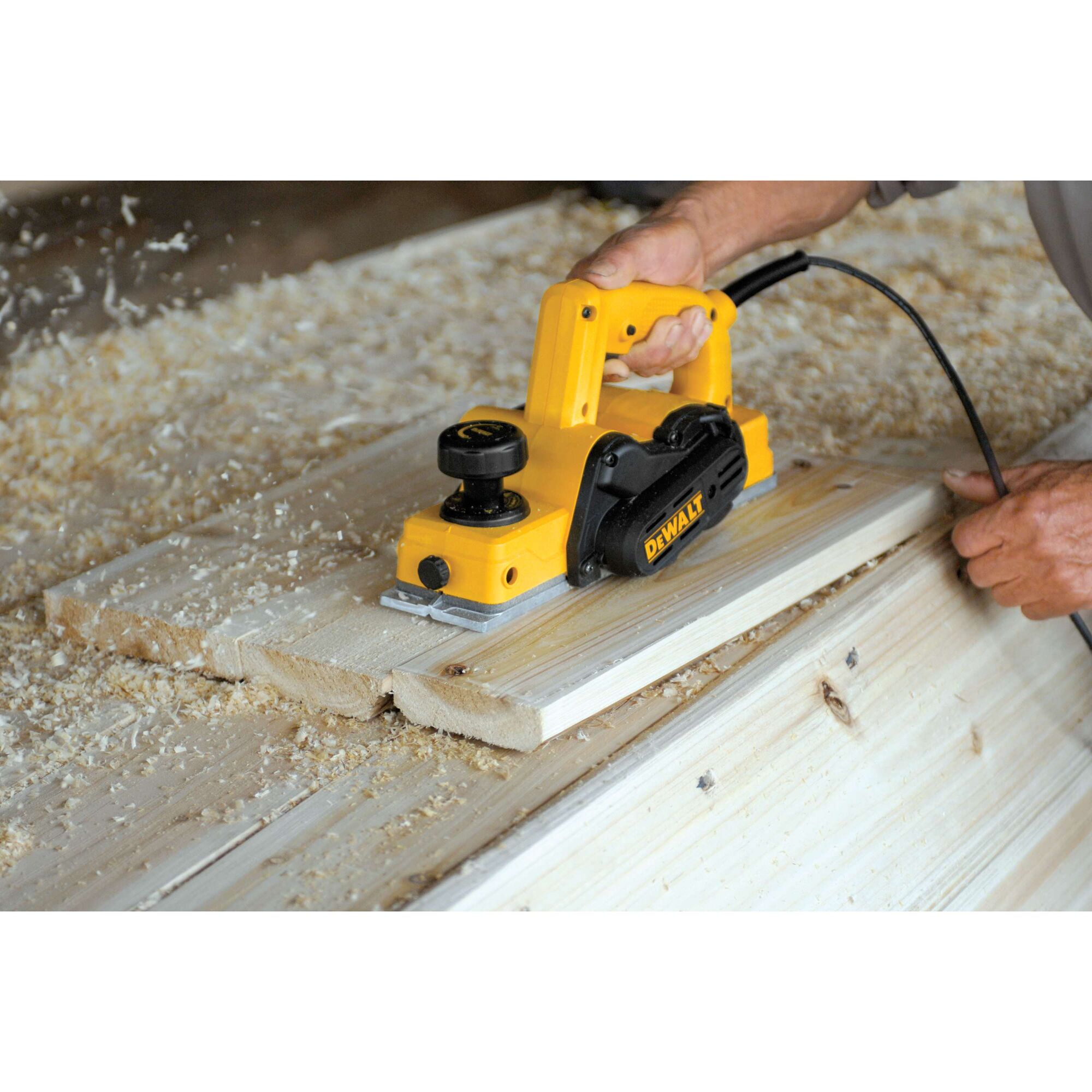 A person using a yellow DEWALT electric hand planer to smooth the surface of wooden boards, with wood shavings scattered around.
