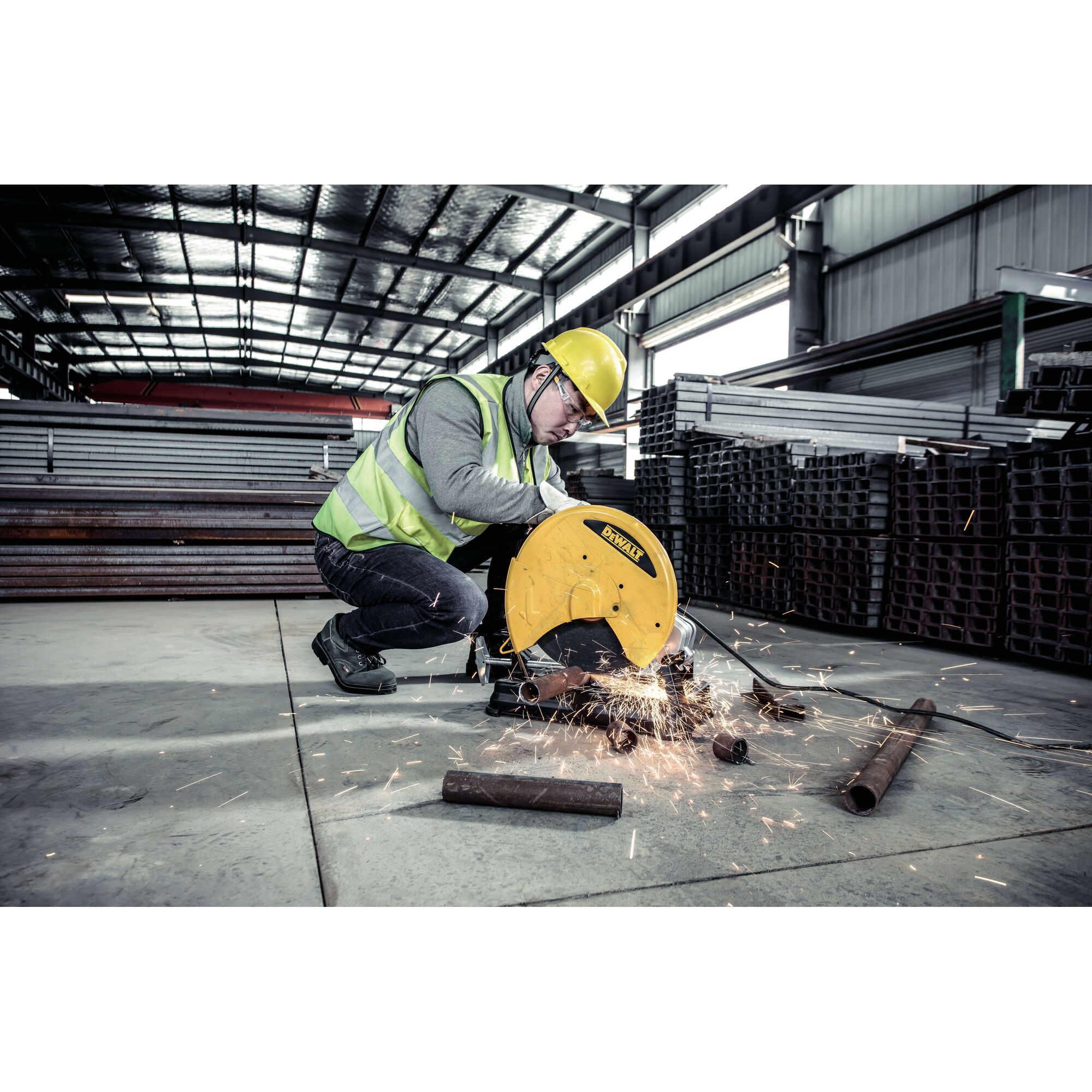 A worker wearing a safety helmet and reflective vest is operating a DEWALT D28730 metal cutting saw, producing sparks as it cuts through metal pipes in an industrial warehouse.