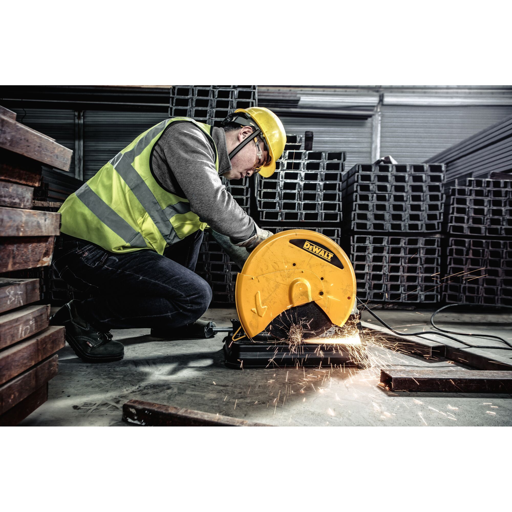 A worker wearing a high-visibility vest and helmet uses a DEWALT D28730 metal cutting chop saw to cut a metal bar, generating sparks in an industrial environment with stacks of metal beams.