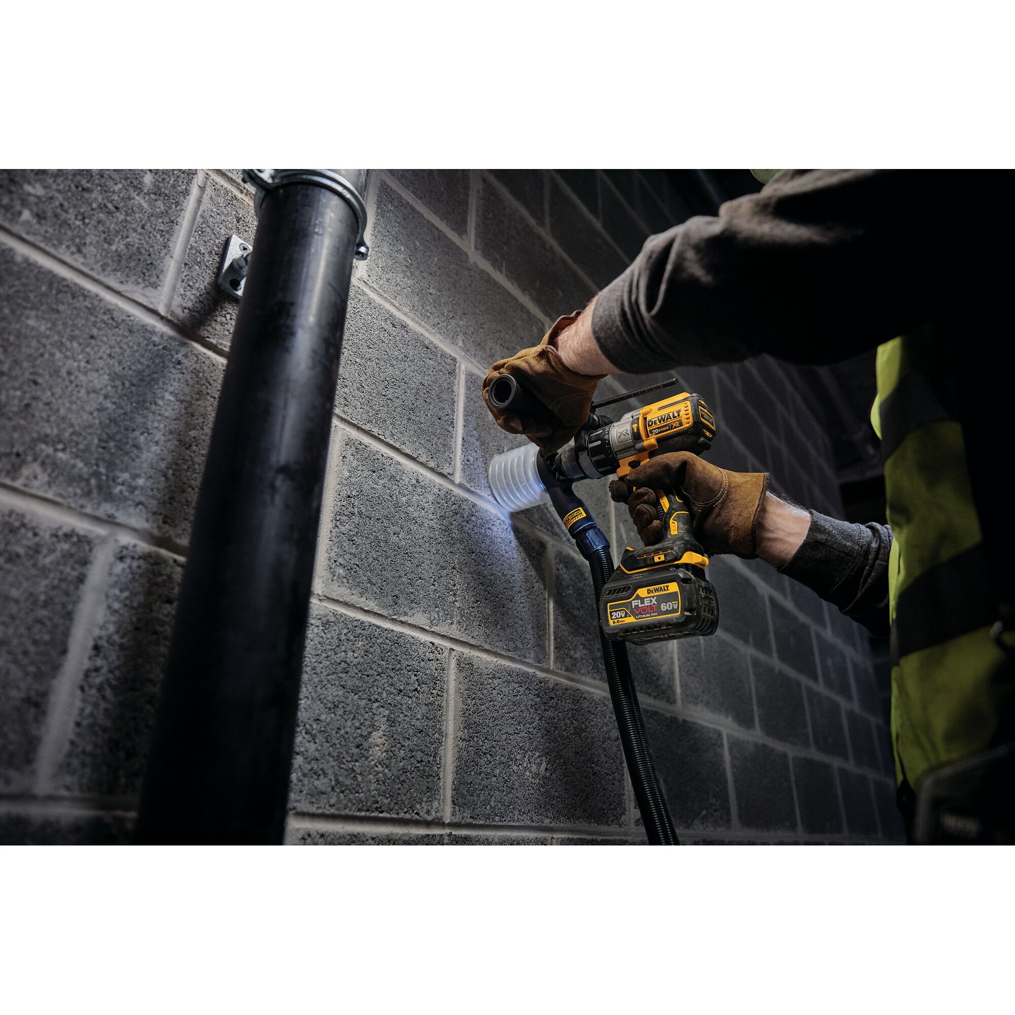 A person wearing work gloves and a safety vest is using a DEWALT cordless drill with a DCB606 FLEXVOLT battery to drill into a concrete block wall, with a dust extraction attachment connected.