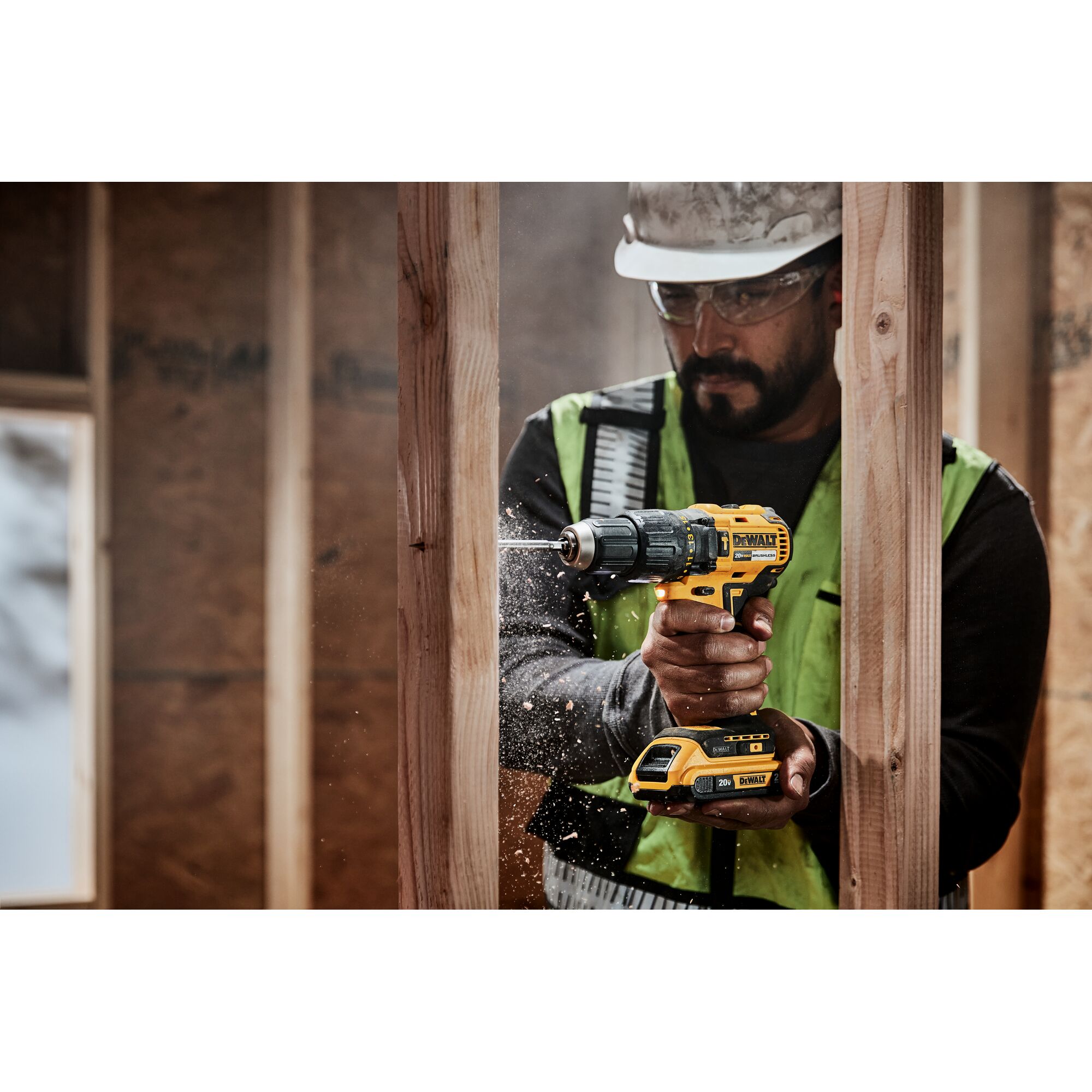A person wearing a safety vest and helmet is using a DEWALT cordless power drill to make a hole in a wooden beam at a construction site. Wood shavings are visible around the drill bit.