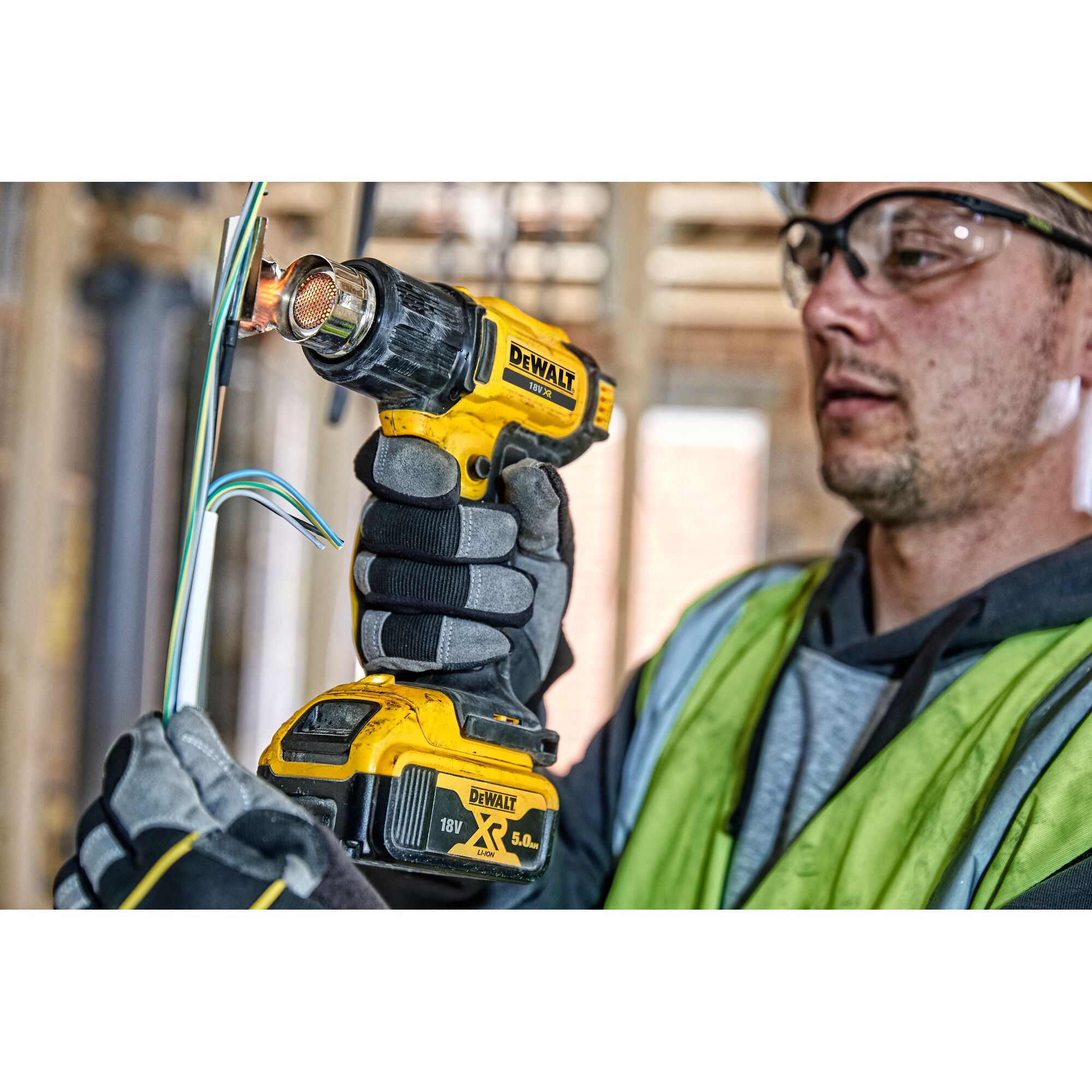 A person wearing gloves and a safety vest is using a DEWALT 18V XR cordless heat gun to work on electrical wires at a construction site. The person's face is blurred.