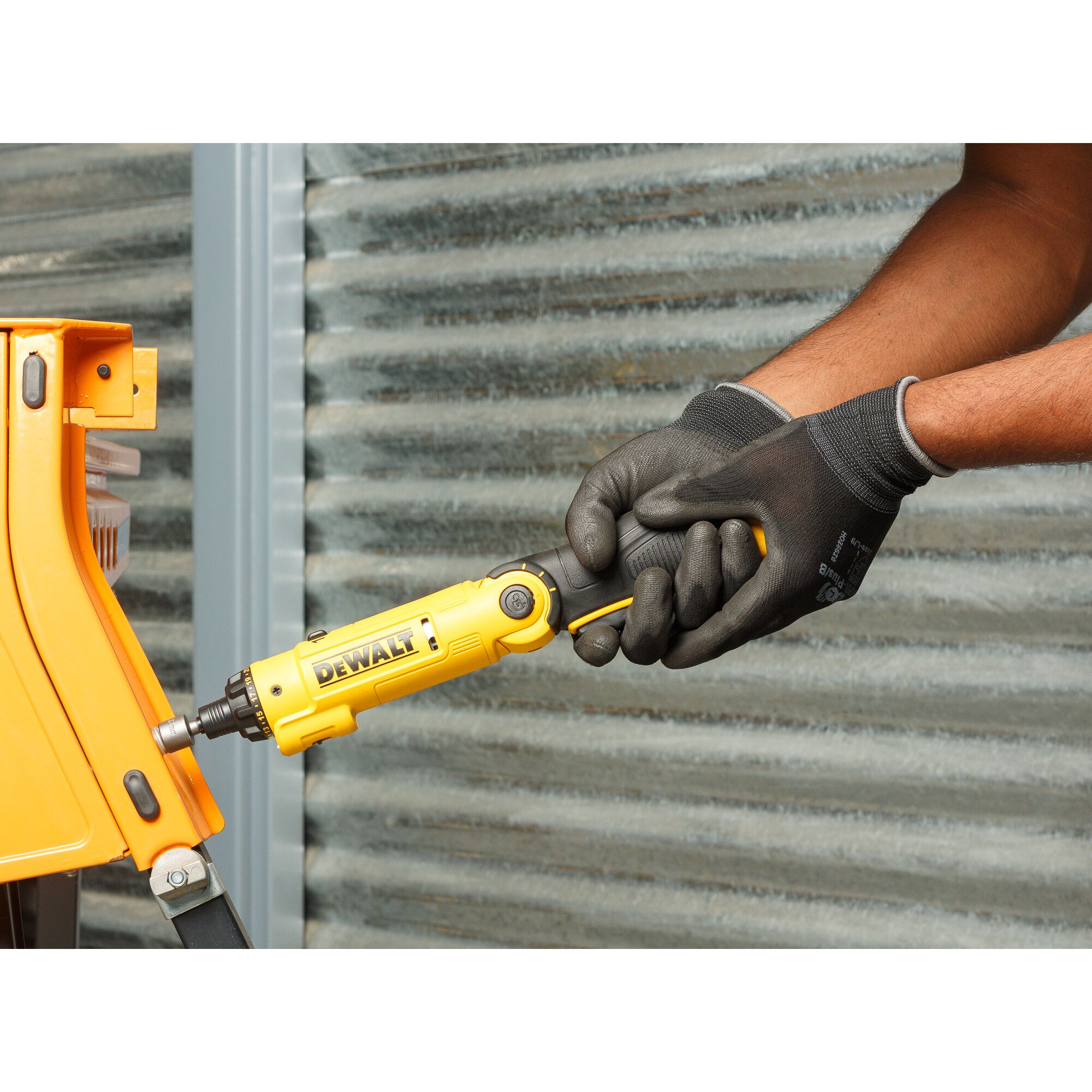 A person wearing black protective gloves is using a yellow DEWALT cordless screwdriver (model DCF008) to fasten a screw on an orange piece of equipment. Corrugated metal wall is visible in the background.