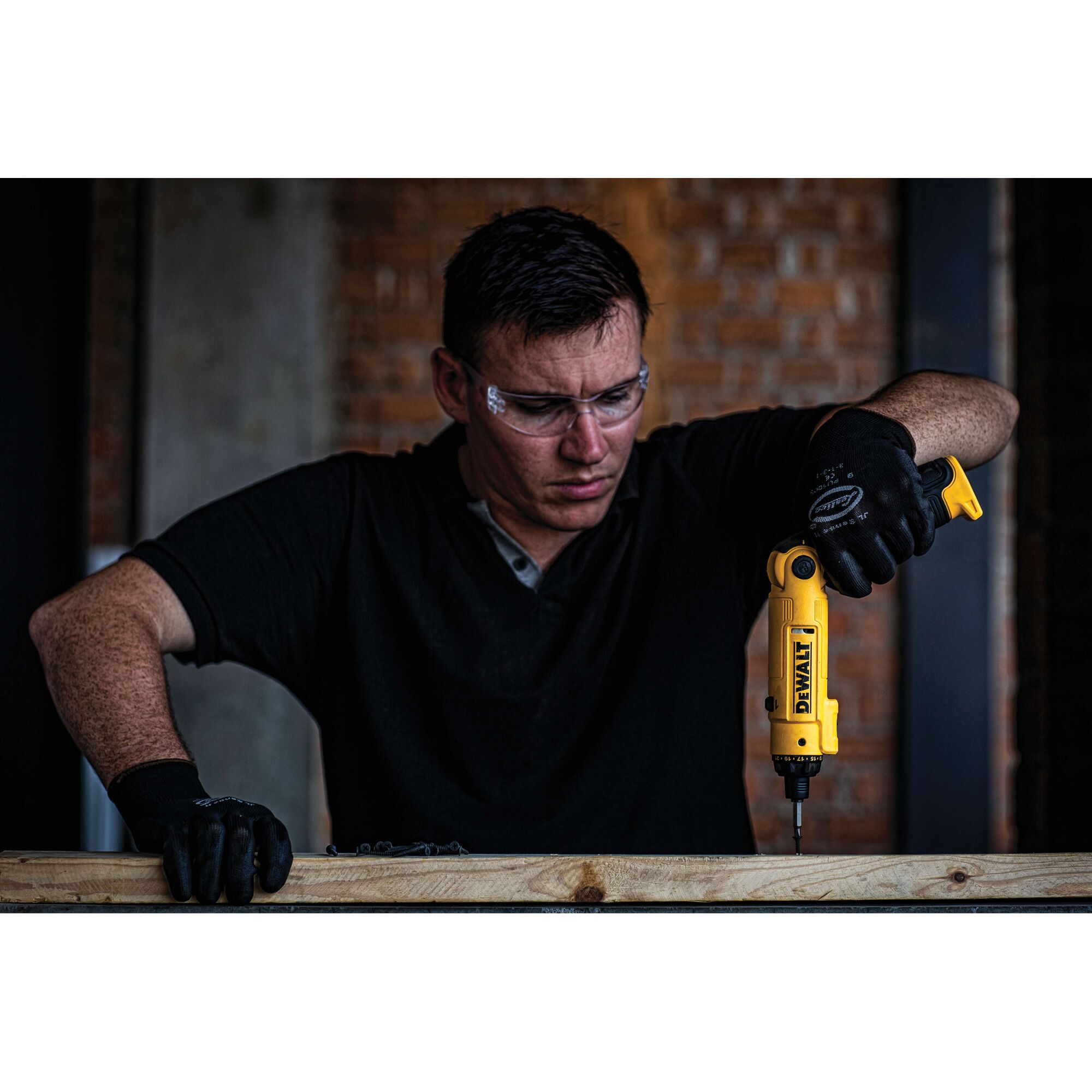 A person wearing black gloves uses a yellow DEWALT DCF008 power tool to drill into a wooden plank. The individual's face is obscured, and the background features a brick wall.