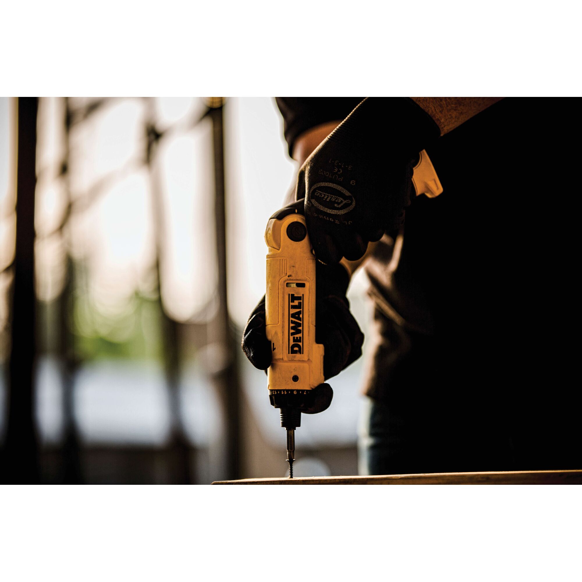 A person wearing black gloves is using a yellow DEWALT cordless screwdriver to drive a screw into a surface. The image is focused on the tool and hand, with a blurred background.