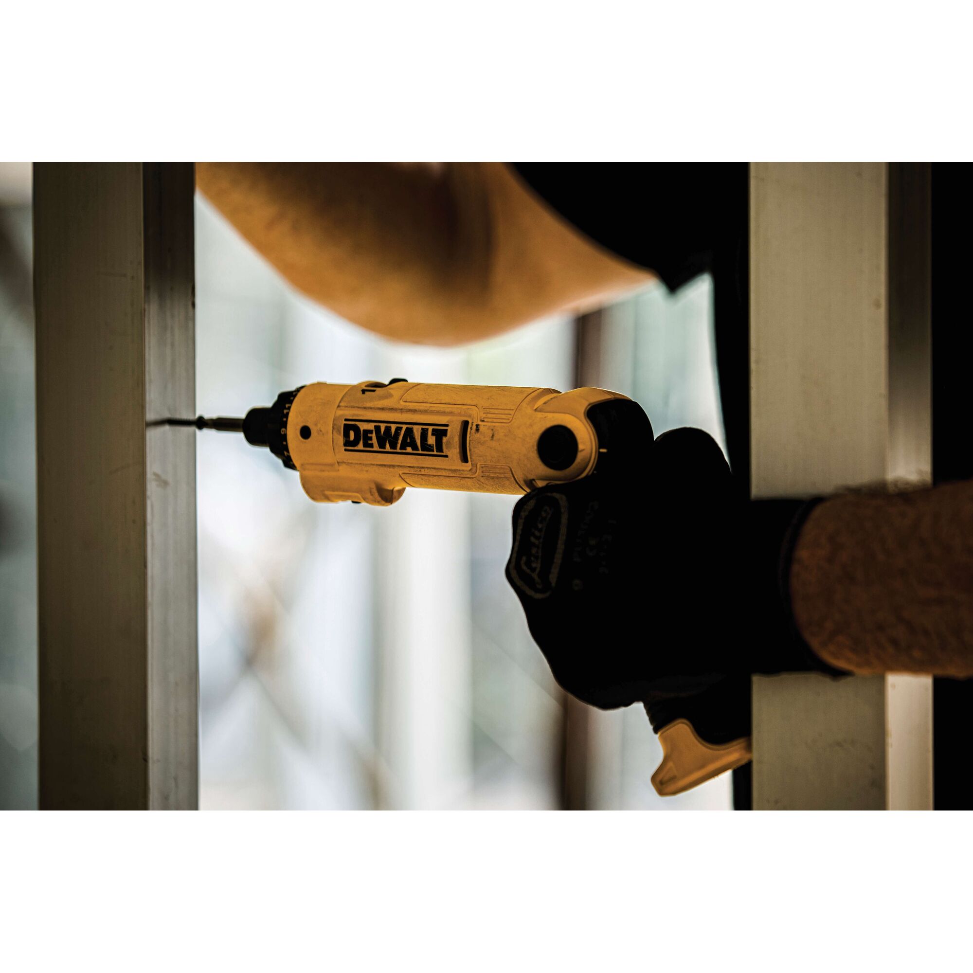 A close-up of a DEWALT electric screwdriver being used to drive a screw into a metal frame. The person is wearing gloves and working indoors.