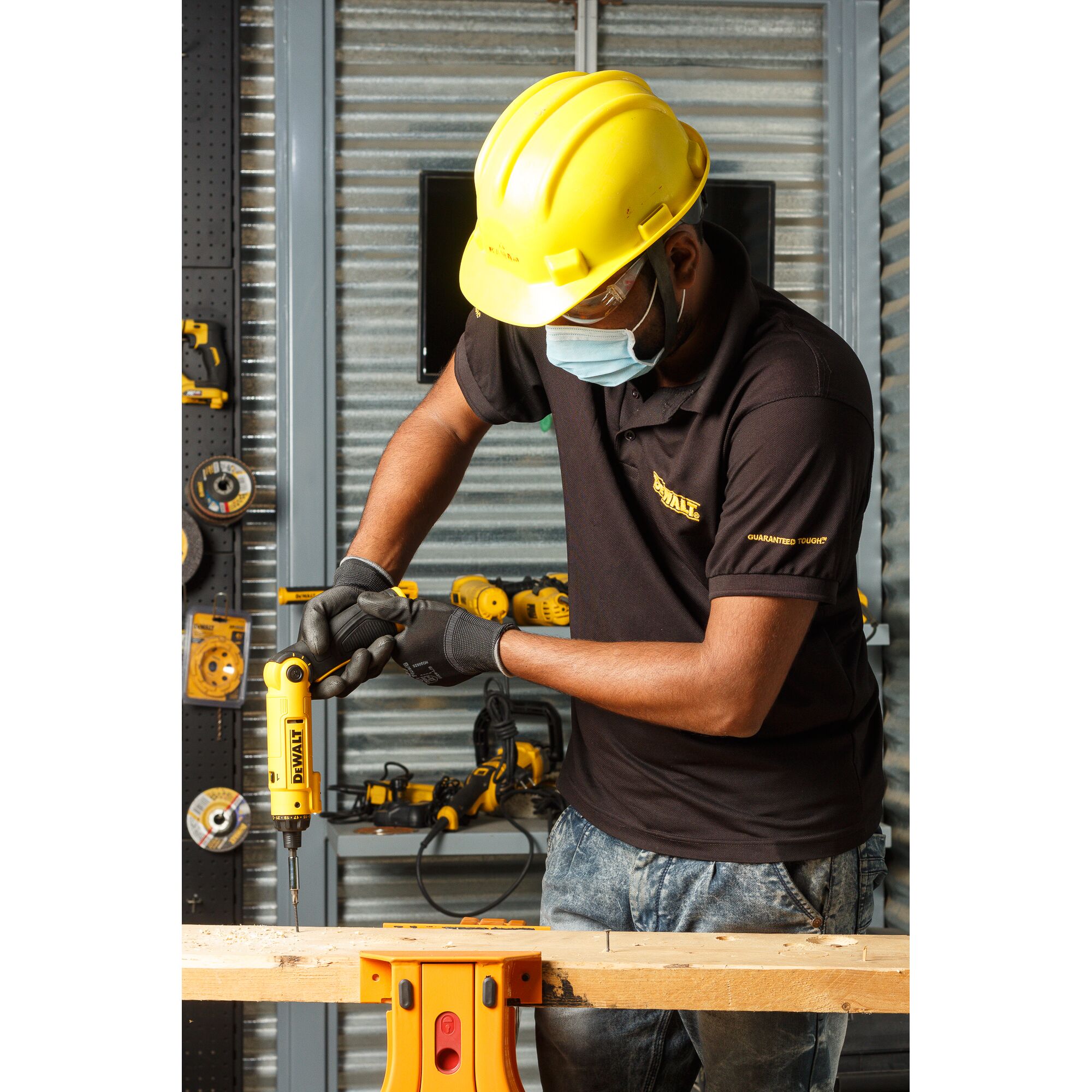 A person wearing a yellow hard hat, black gloves, and a black DEWALT shirt is using a yellow DEWALT DCF008 power tool to drill into a piece of wood in a workshop setting.