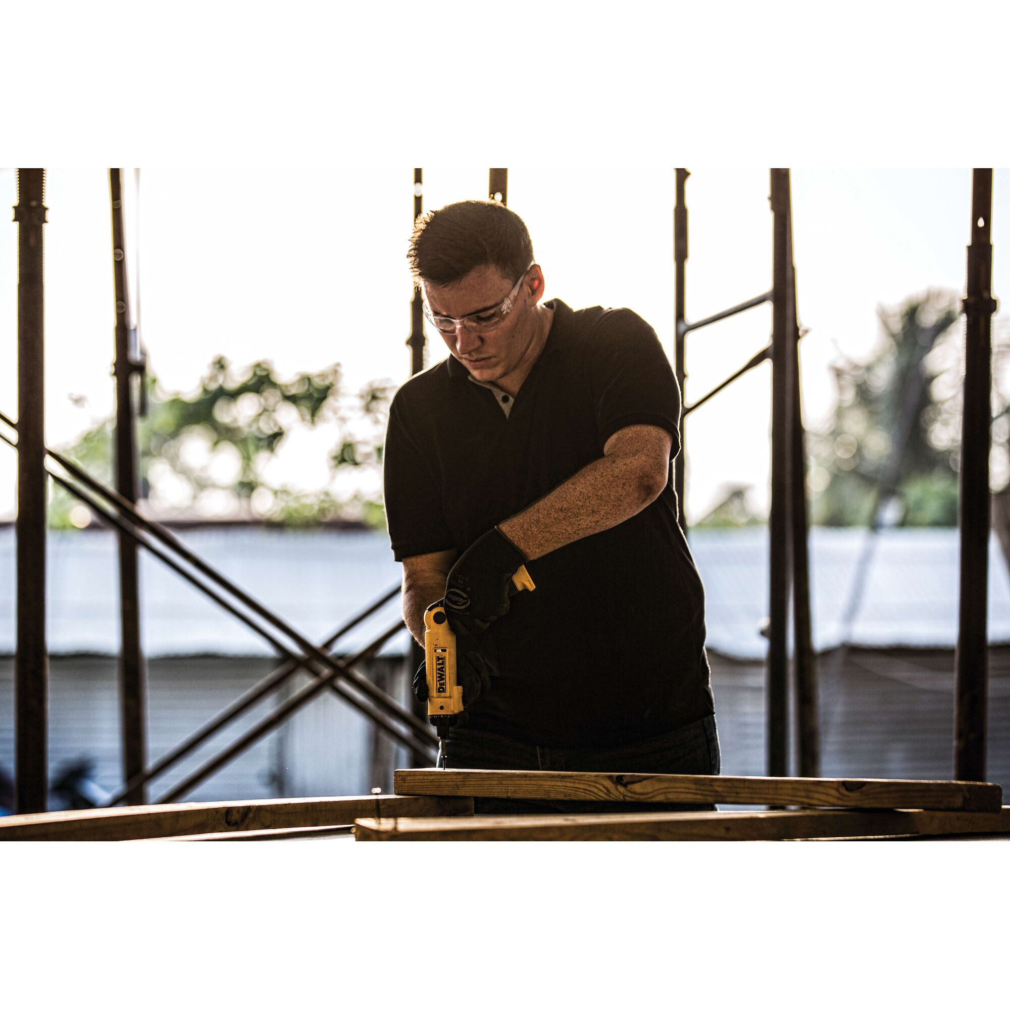 A person wearing black gloves operates a yellow DEWALT DCF008 power tool to drill into a piece of wood at a construction site.