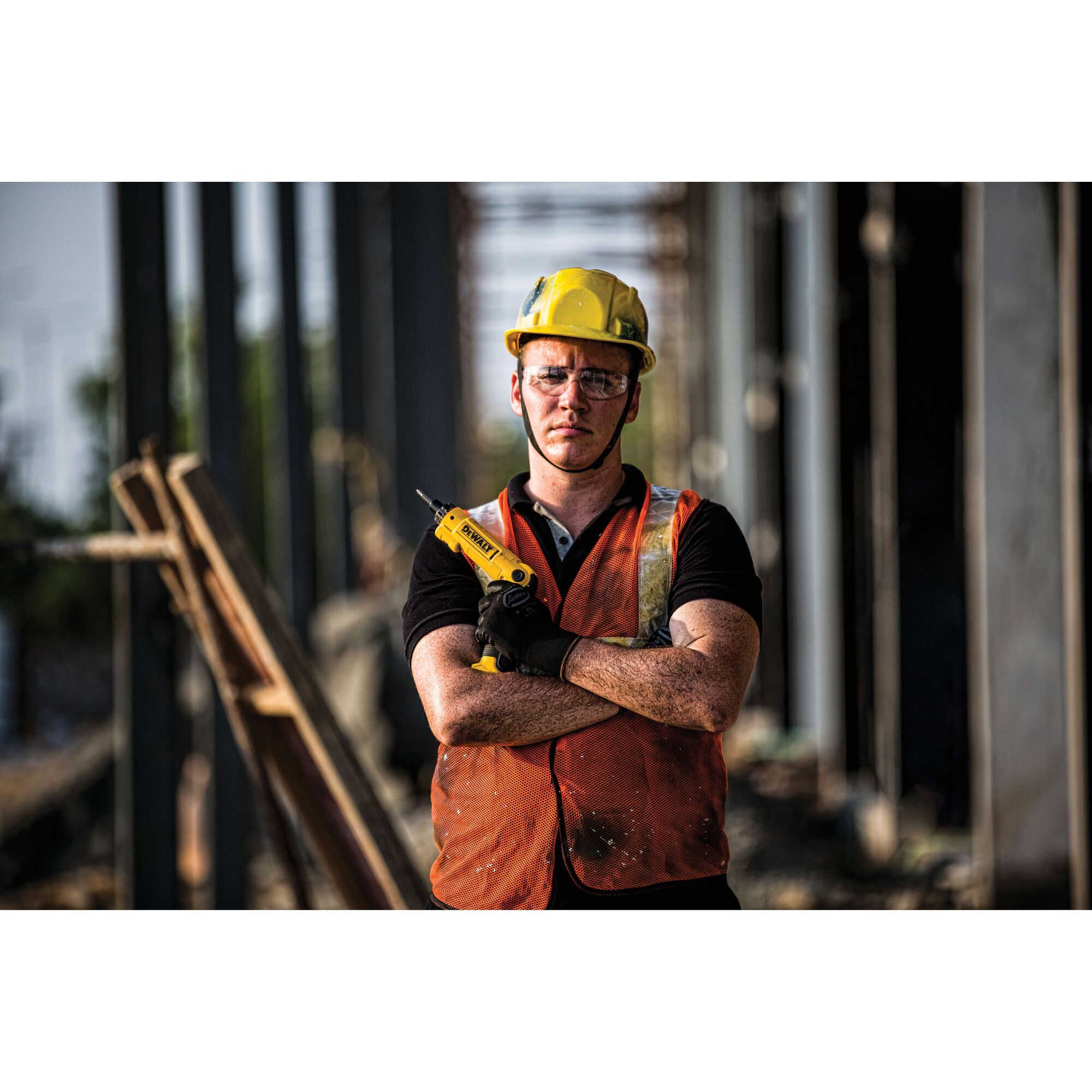 A construction worker wearing a yellow hard hat and orange safety vest stands with arms crossed, holding a DEWALT DCF008 tool in one hand at a building site. The worker's face is blurred for privacy.