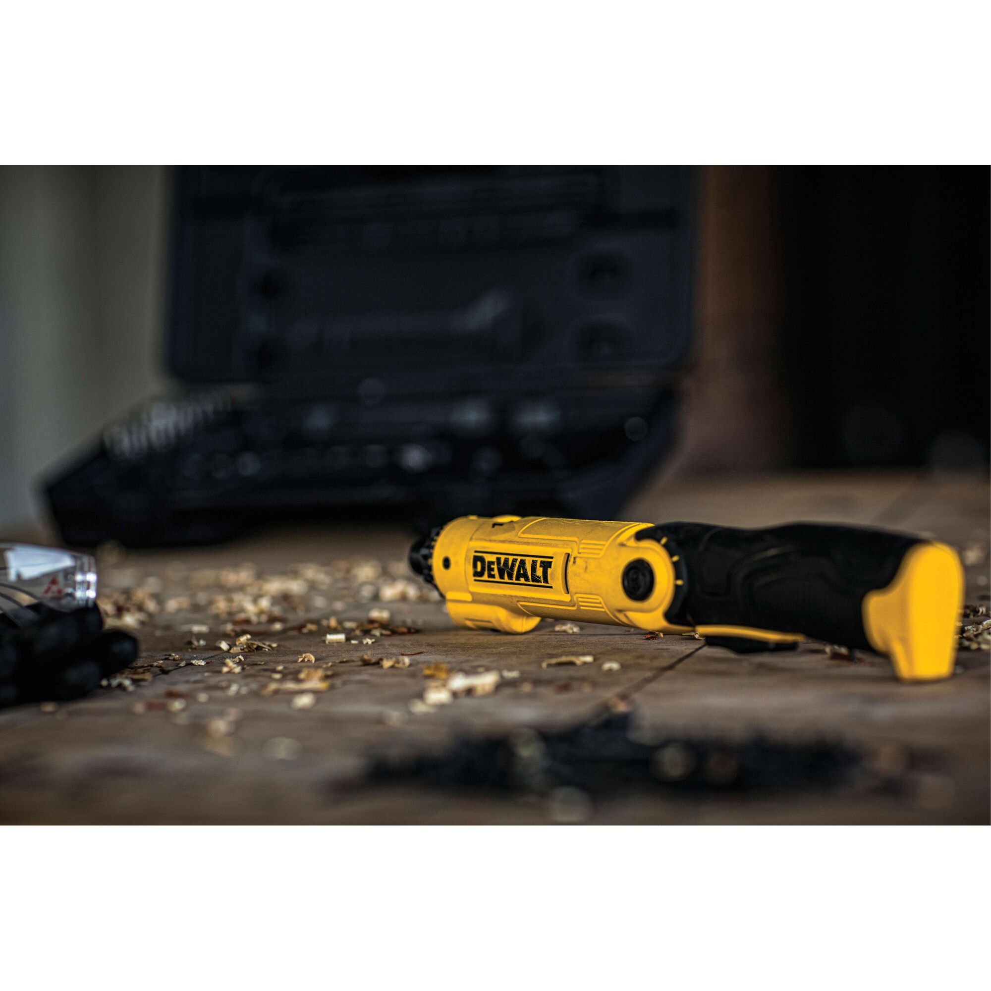A yellow and black DEWALT cordless screwdriver (model DCF008) lying on a wooden surface with wood shavings, with a black case and other tools blurred in the background.