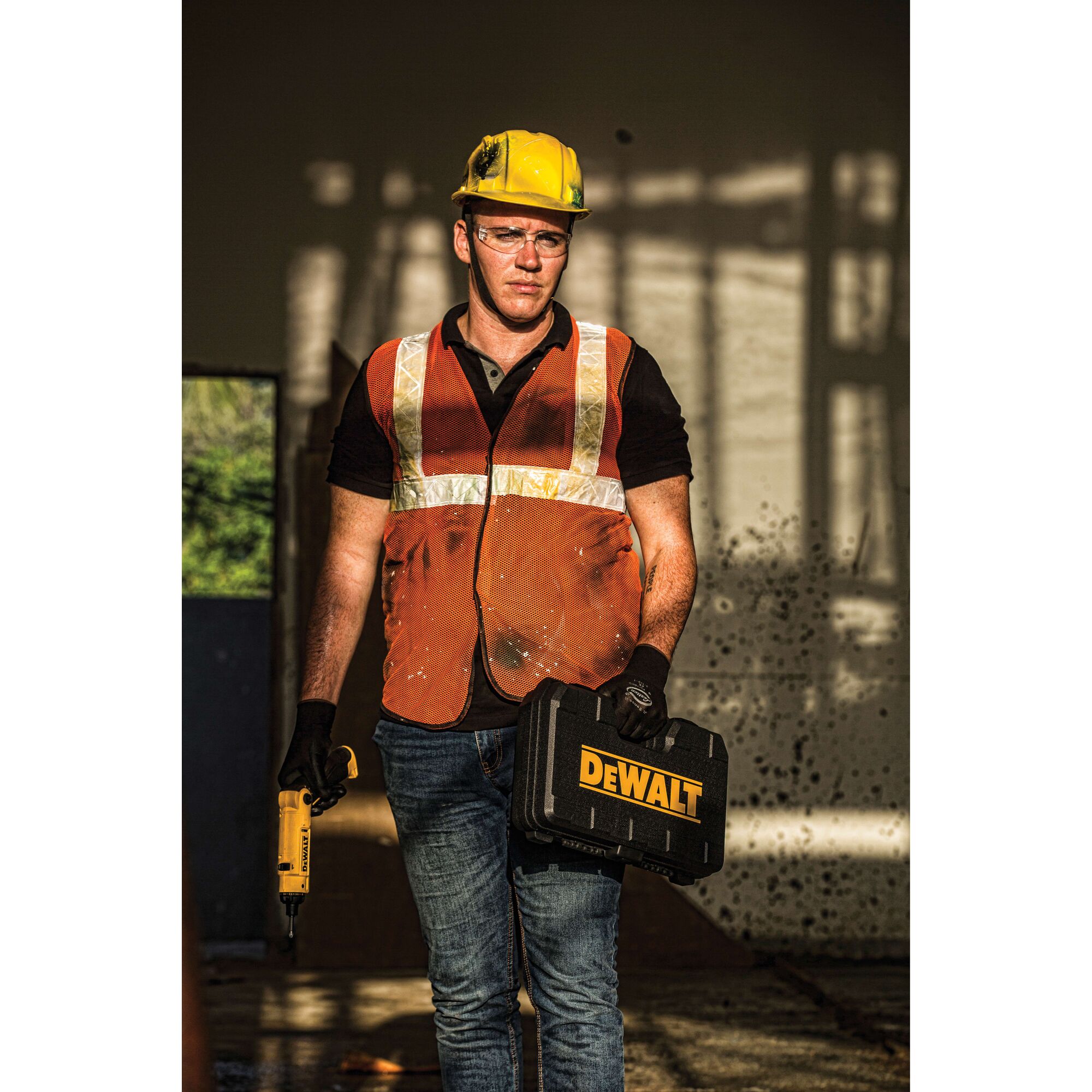 A person wearing a yellow safety helmet, an orange reflective vest, and gloves stands indoors holding a DEWALT-branded black toolbox in one hand and a yellow DEWALT power tool in the other hand. The background is a construction or industrial setting.