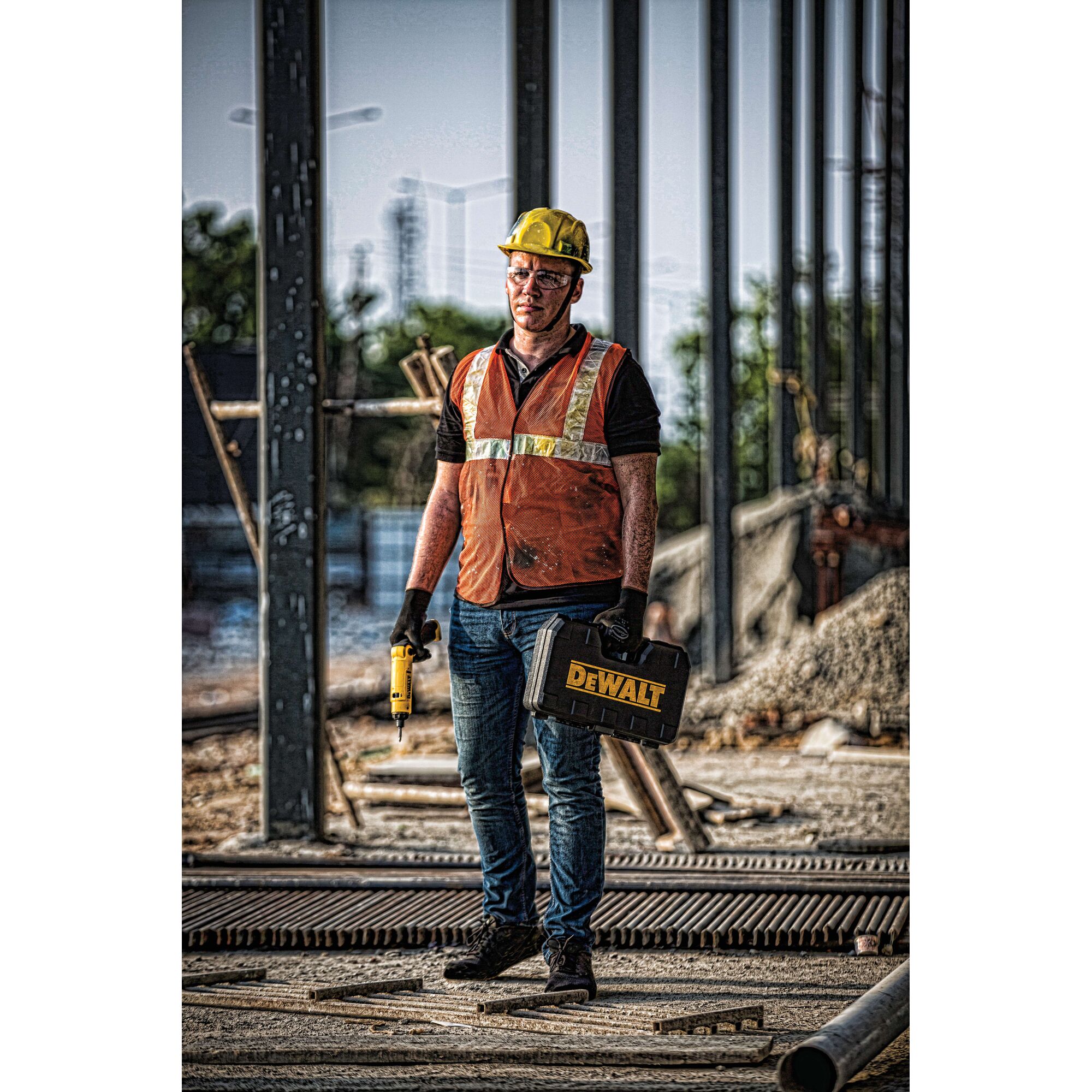 A construction worker wearing a yellow hard hat and orange safety vest stands at a construction site, holding a DEWALT toolbox in one hand and a cordless tool in the other.
