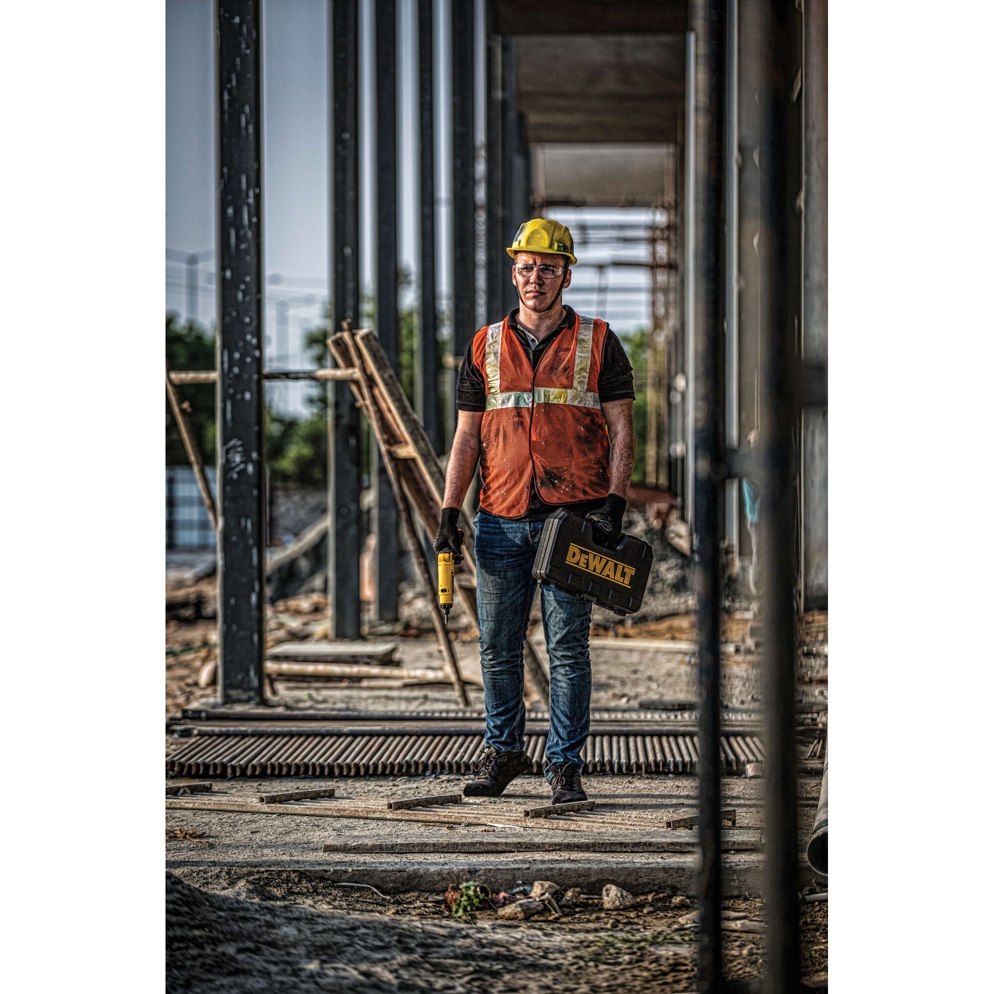 A construction worker wearing a yellow hard hat and orange safety vest stands on a building site holding a DEWALT toolbox and power tool. The worker's face is blurred for privacy, and the area has steel beams and construction materials.