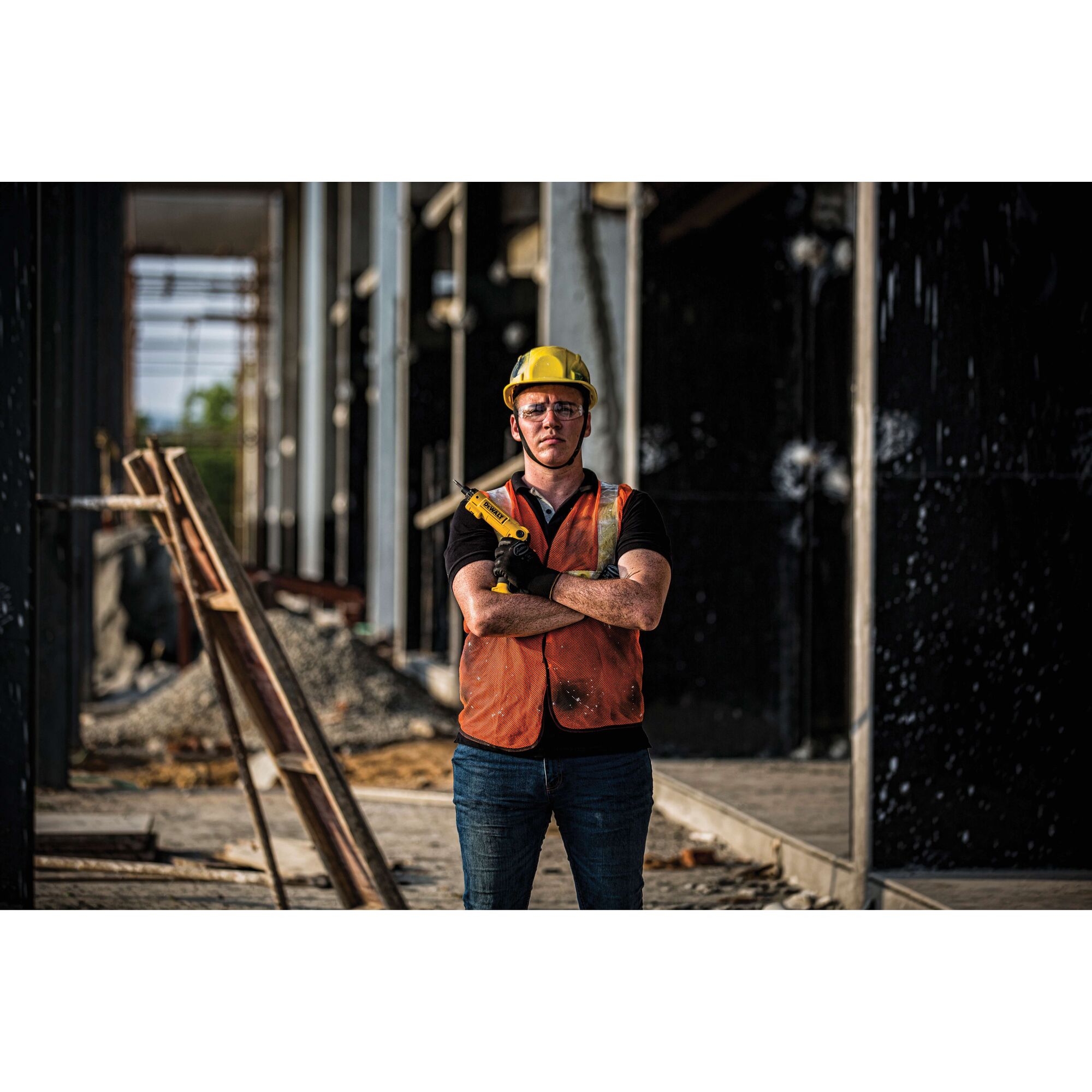 A person wearing a yellow safety helmet and orange vest stands with arms crossed at a construction site, holding a DEWALT tool. The background shows building framework and construction materials.