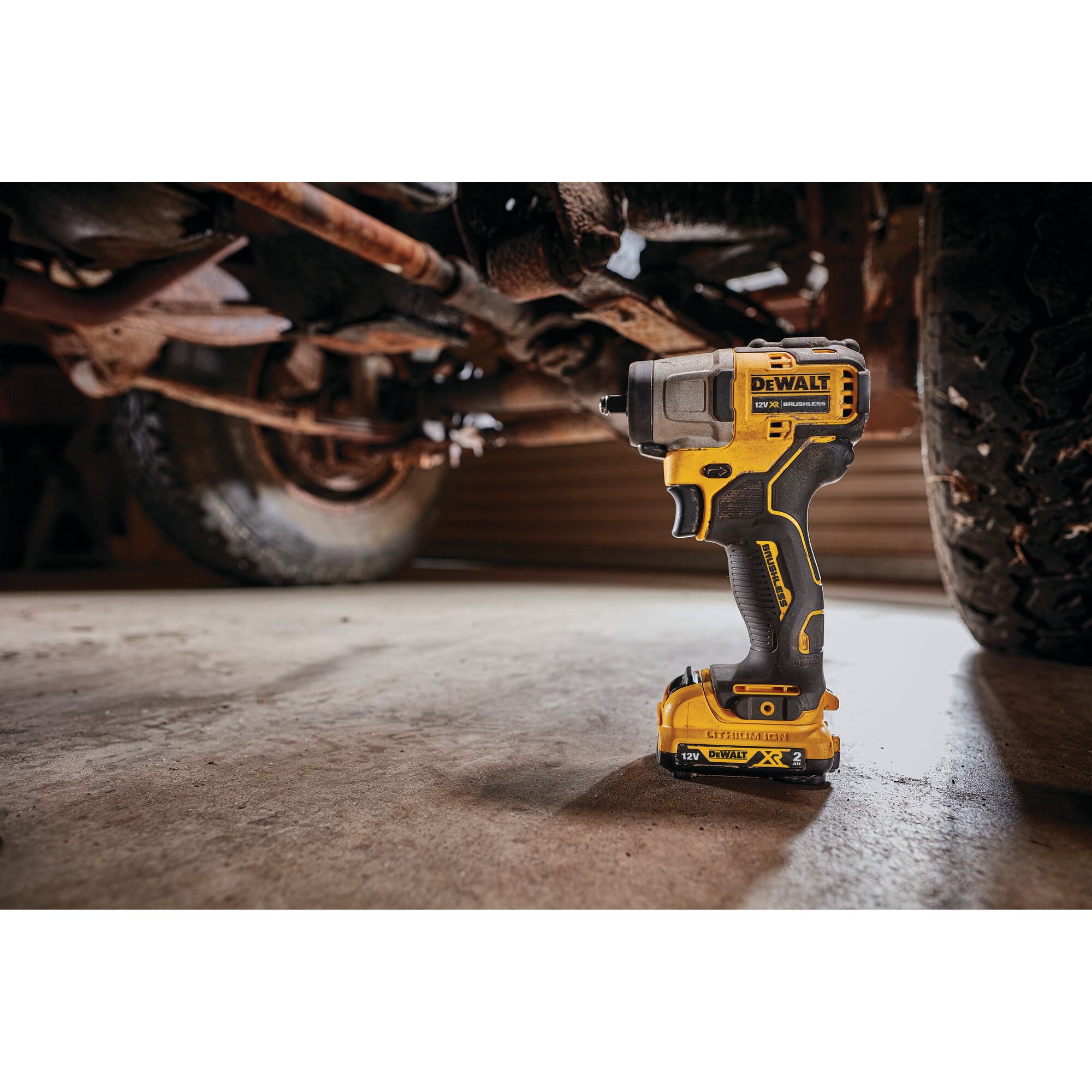 A DEWALT DCF902D2 cordless impact wrench with a yellow and black design is shown standing upright on a garage floor, positioned beneath a vehicle with visible suspension and tire components.