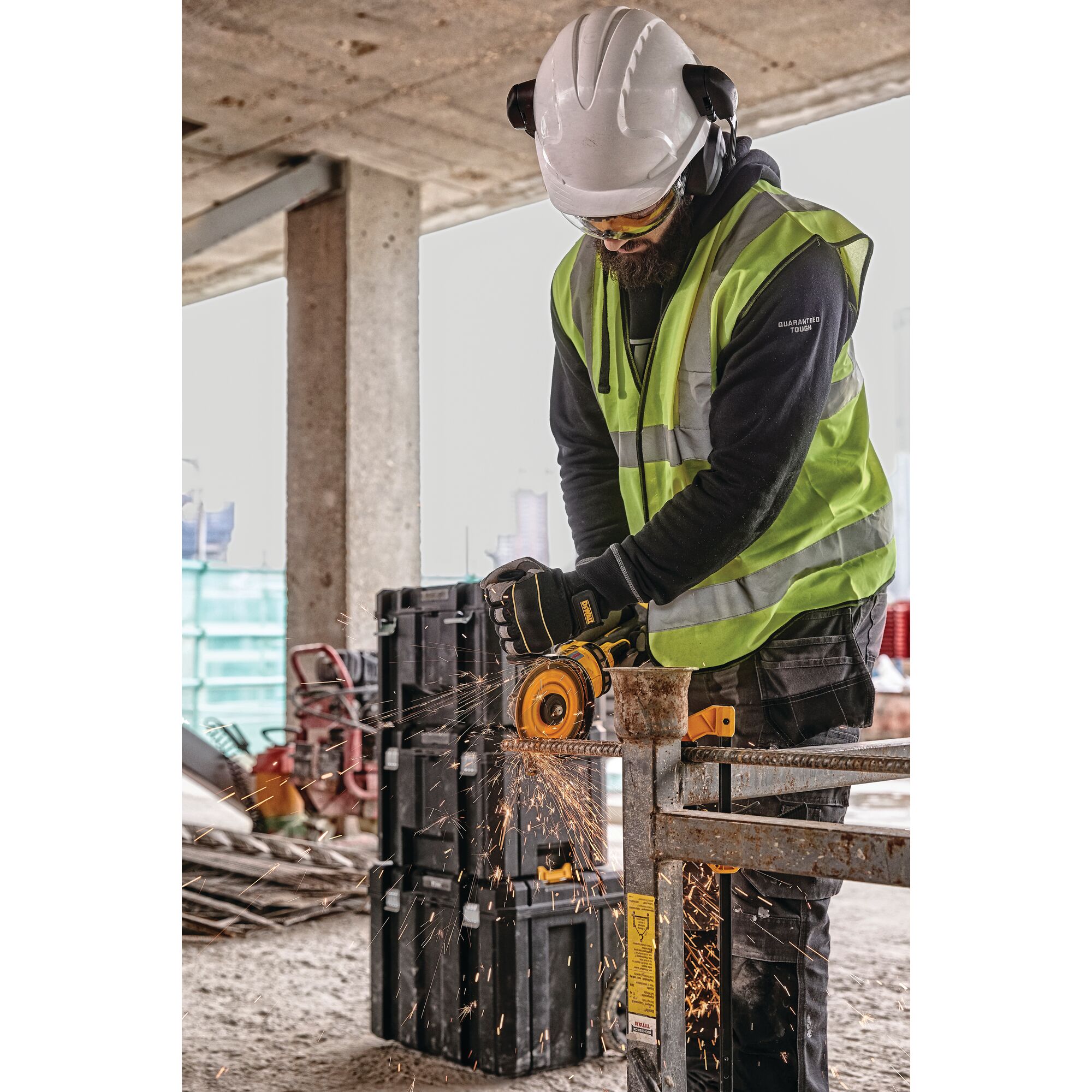 A construction worker wearing a white safety helmet, safety glasses, ear protection, gloves, and a high visibility vest is using a yellow DEWALT angle grinder to cut through a metal bar, producing sparks, at a construction site.