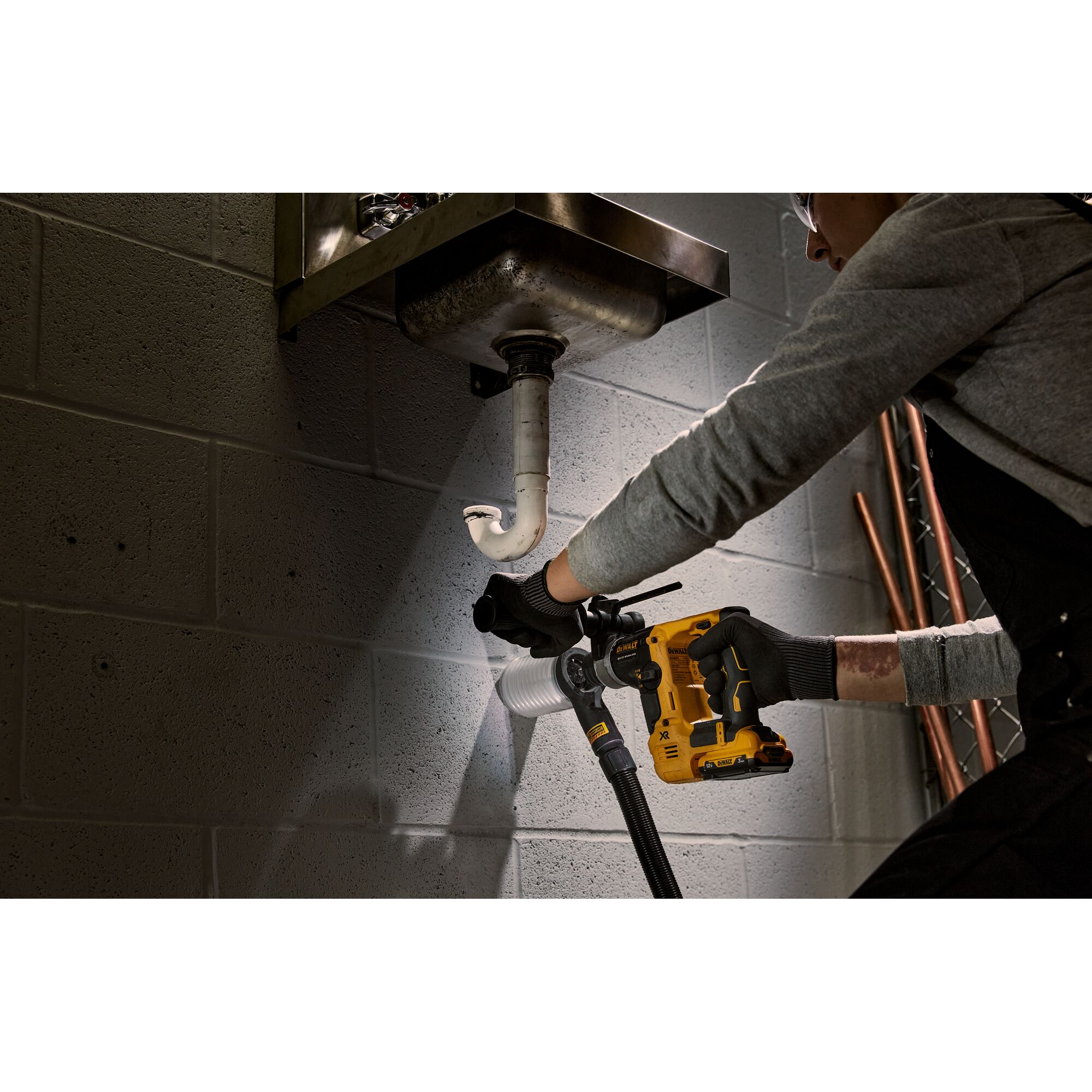 A person wearing gloves uses a DEWALT yellow cordless rotary hammer drill under a sink to work on pipes attached to a concrete wall. The drill is model DCH072L2 and is being used in a plumbing setting.