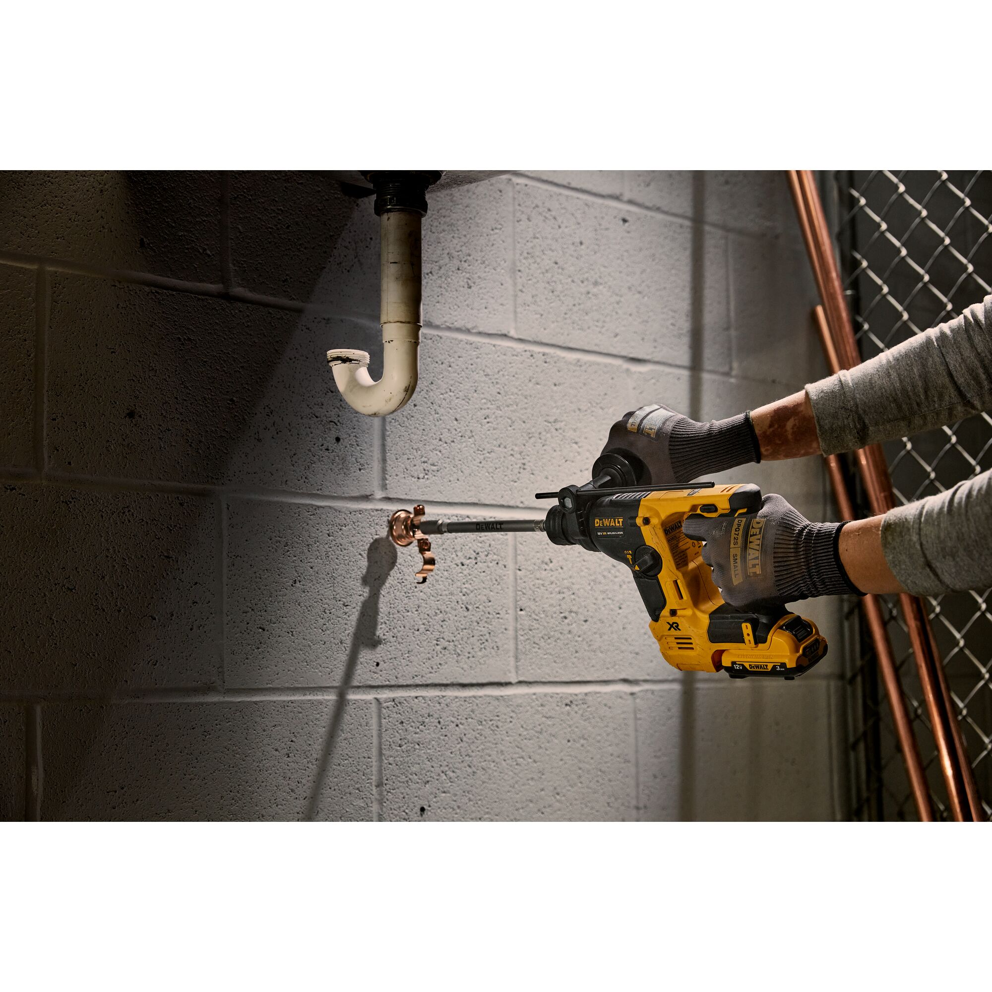 A person using a DEWALT cordless rotary hammer drill to drill into a concrete wall beneath plumbing pipes. The person is wearing gloves and the tool is yellow and black.