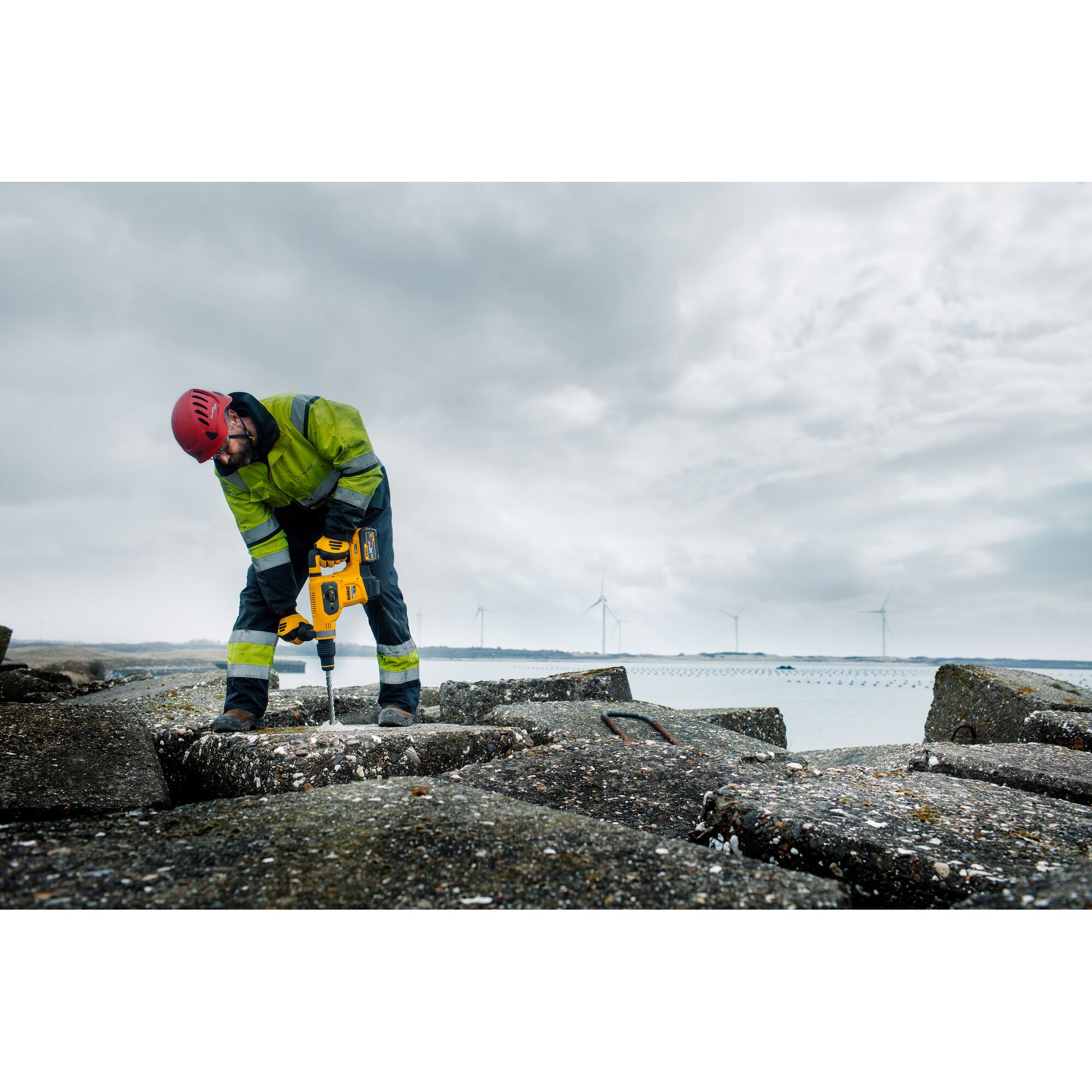 A construction worker wearing a red helmet and green high-visibility jacket is using a yellow DEWALT rotary hammer drill (DCH481N) on large concrete blocks outdoors. Wind turbines are visible in the background under a cloudy sky.