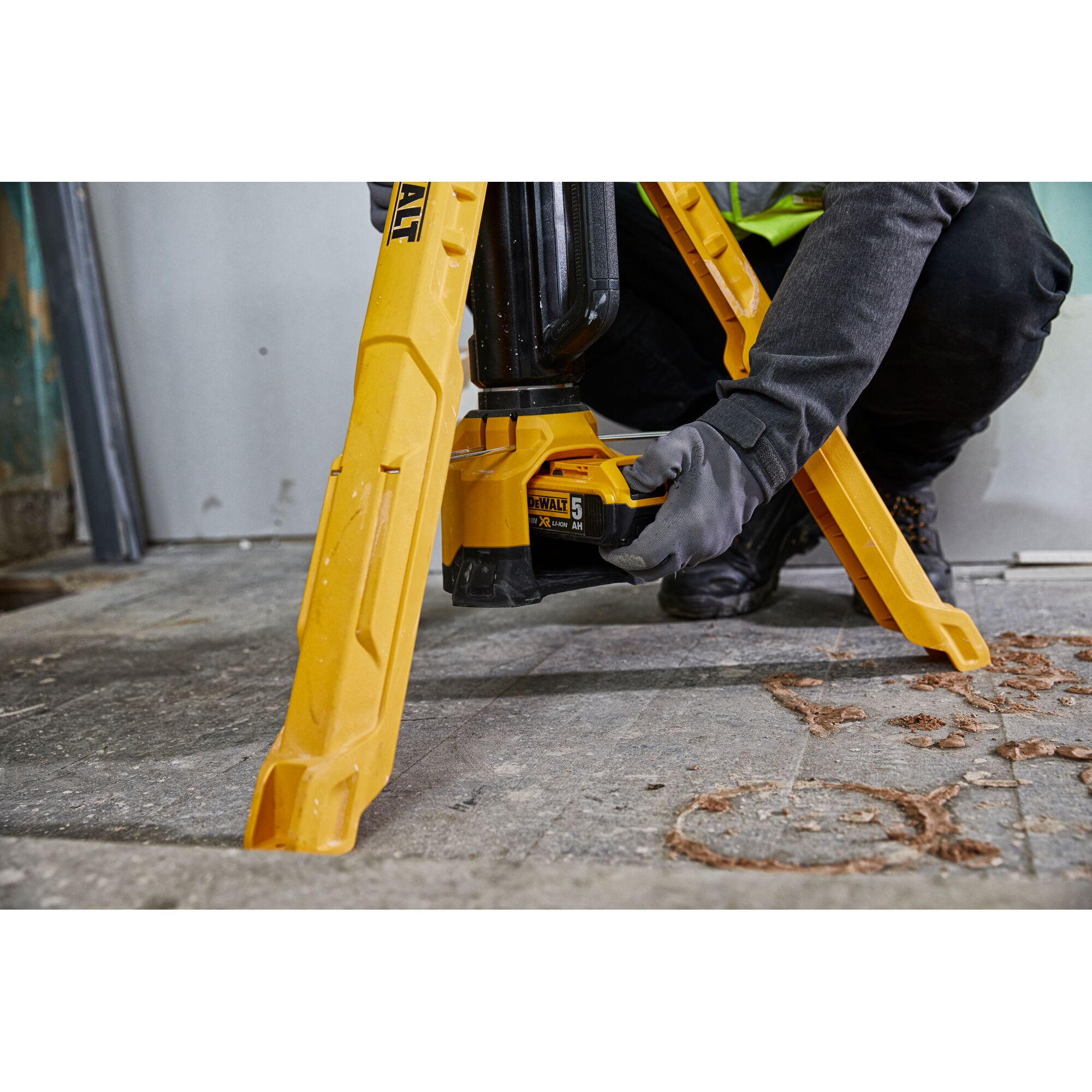 Person installs battery in a yellow DEWALT tripod work light on a workshop floor.