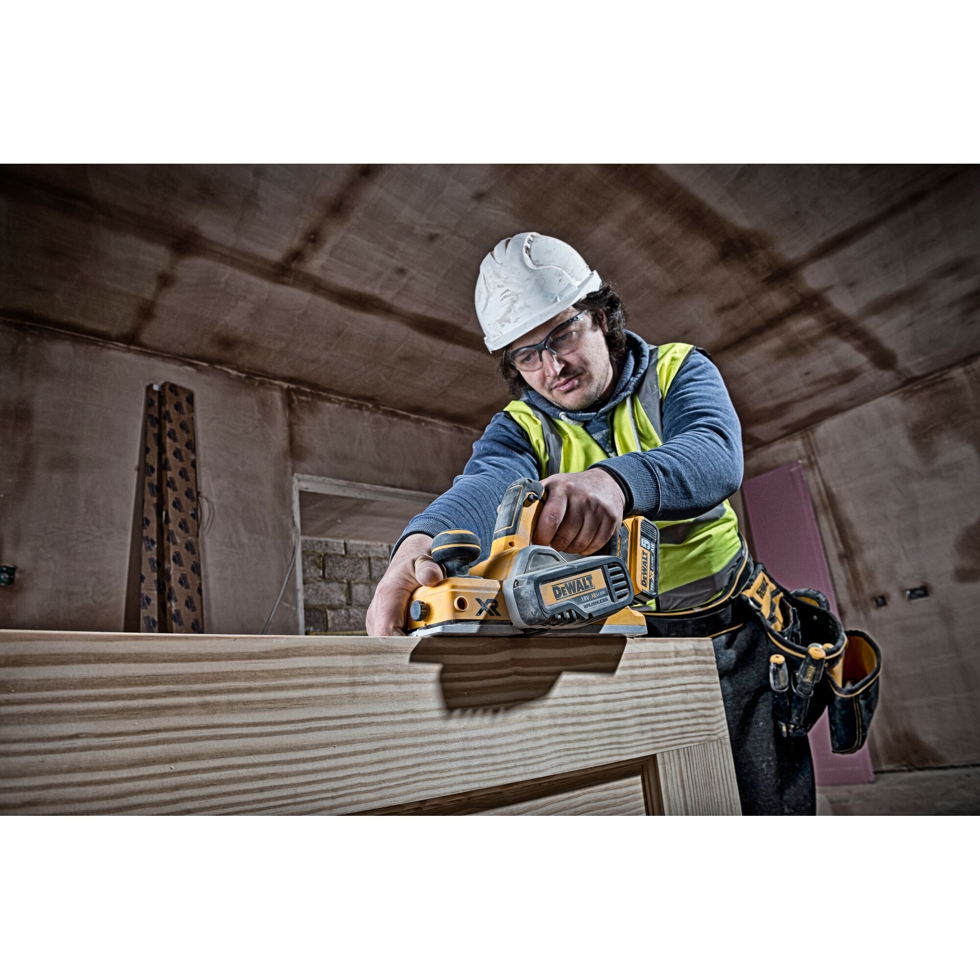 Worker uses DEWALT DCP580N planer on wood indoors, wearing safety gear and tool belt.