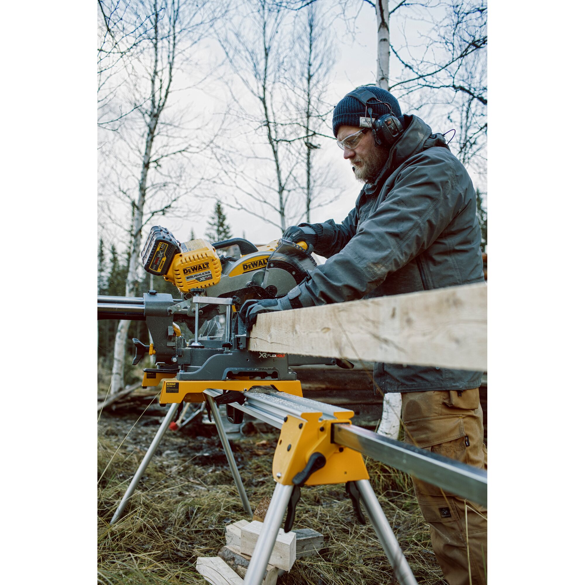A person using a DEWALT DHS780T2 miter saw on a stand outdoors to cut a long wooden board. The person is wearing protective gloves, earmuffs, and a jacket. Leafless trees are visible in the background.