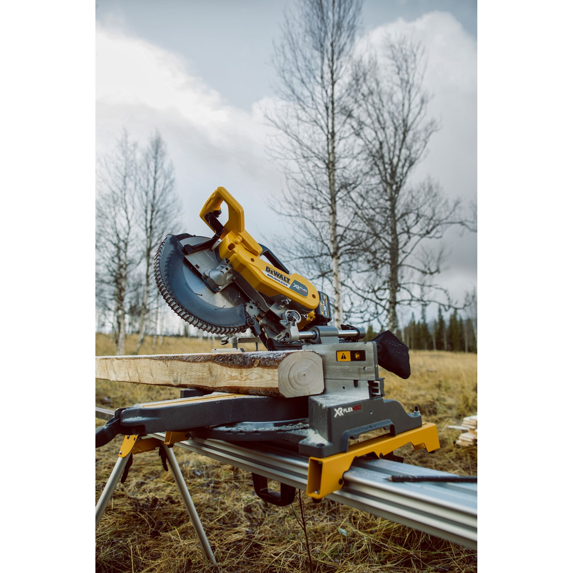 DEWALT miter saw model DHS780T2 set up outdoors on a workbench, cutting a wooden beam. Leafless trees and a cloudy sky are visible in the background.