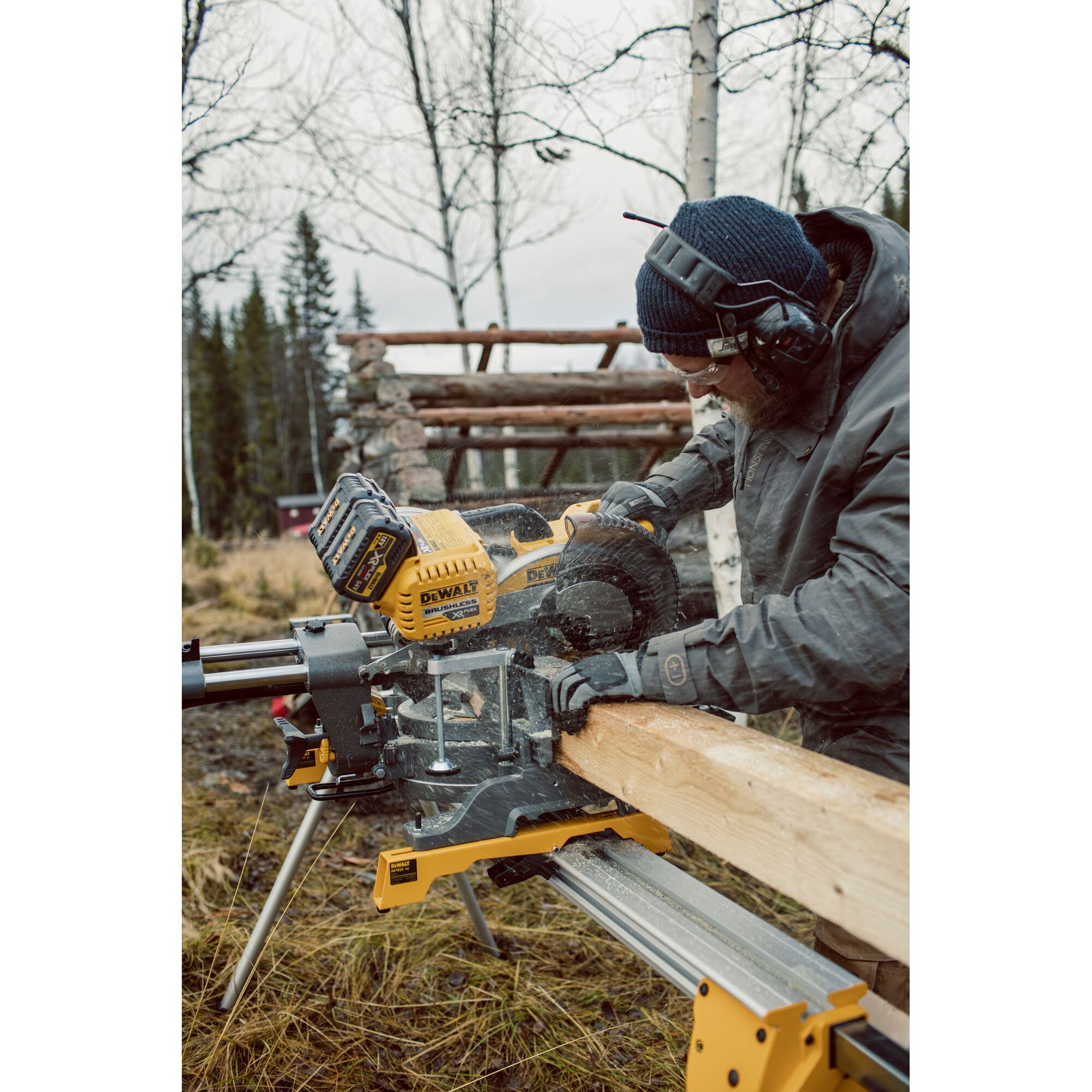 A person using a DEWALT DHS780T2 miter saw to cut a wooden beam outdoors, wearing safety earmuffs, gloves, and a beanie. The saw is set up on a stand in a woodland area.