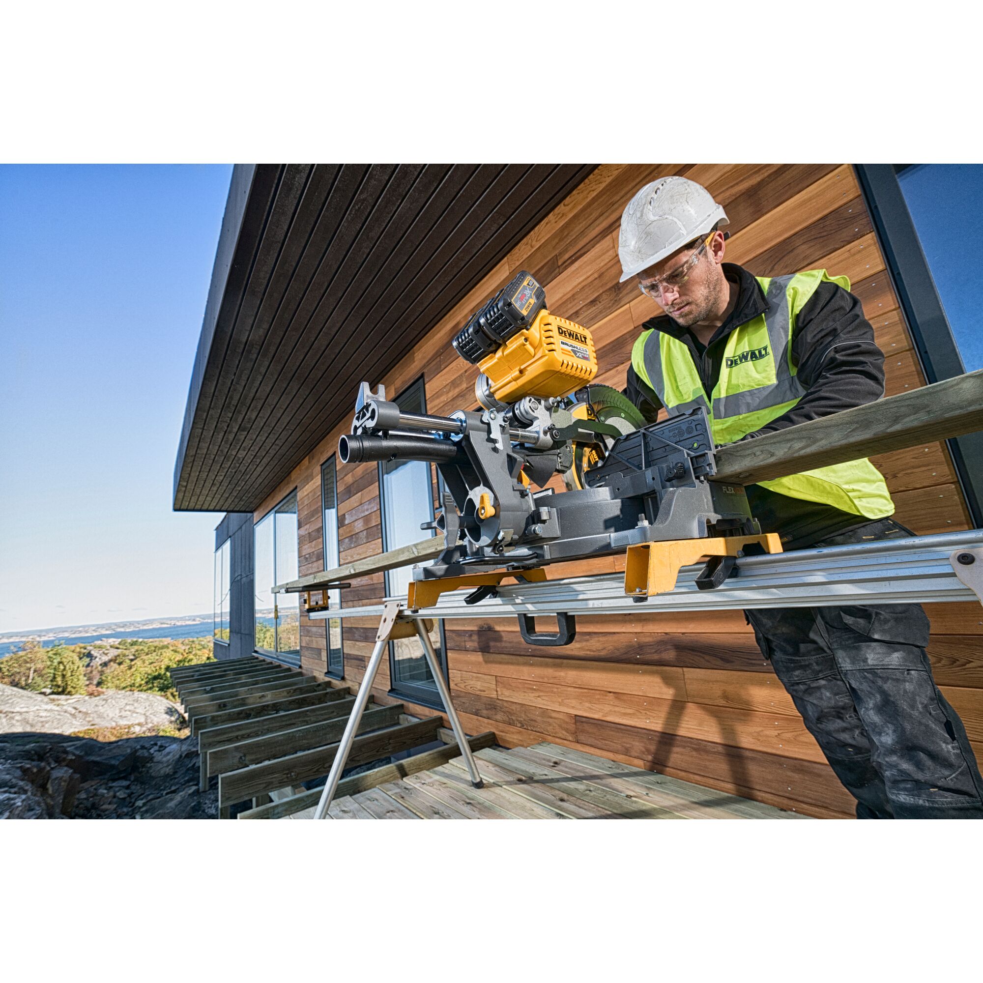 A person wearing a safety helmet and a high-visibility vest is using a DEWALT DHS780T2 miter saw to cut a wooden beam outdoors, next to a modern wooden building under construction.