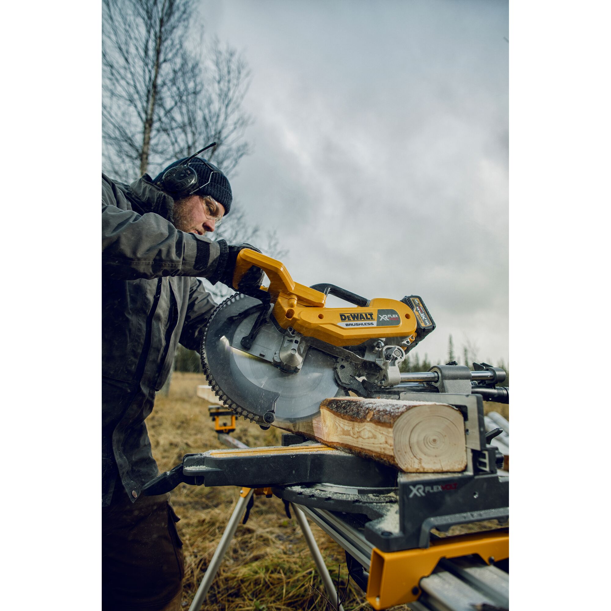 A person using a DEWALT DHS780T2 XR FLEXVOLT cordless saw to cut a log outdoors, with protective clothing and ear protection, set on a portable saw stand.