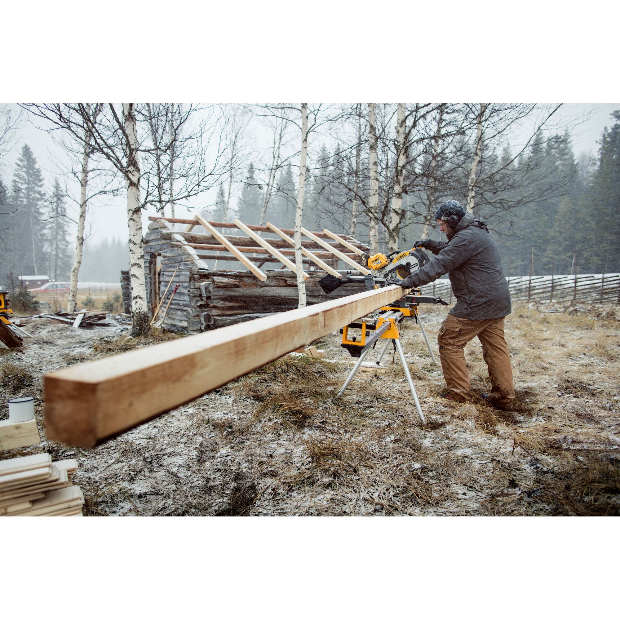 A person using a DEWALT sliding compound miter saw outdoors to cut a long wooden beam, with a partially built wooden structure and trees in the background.
