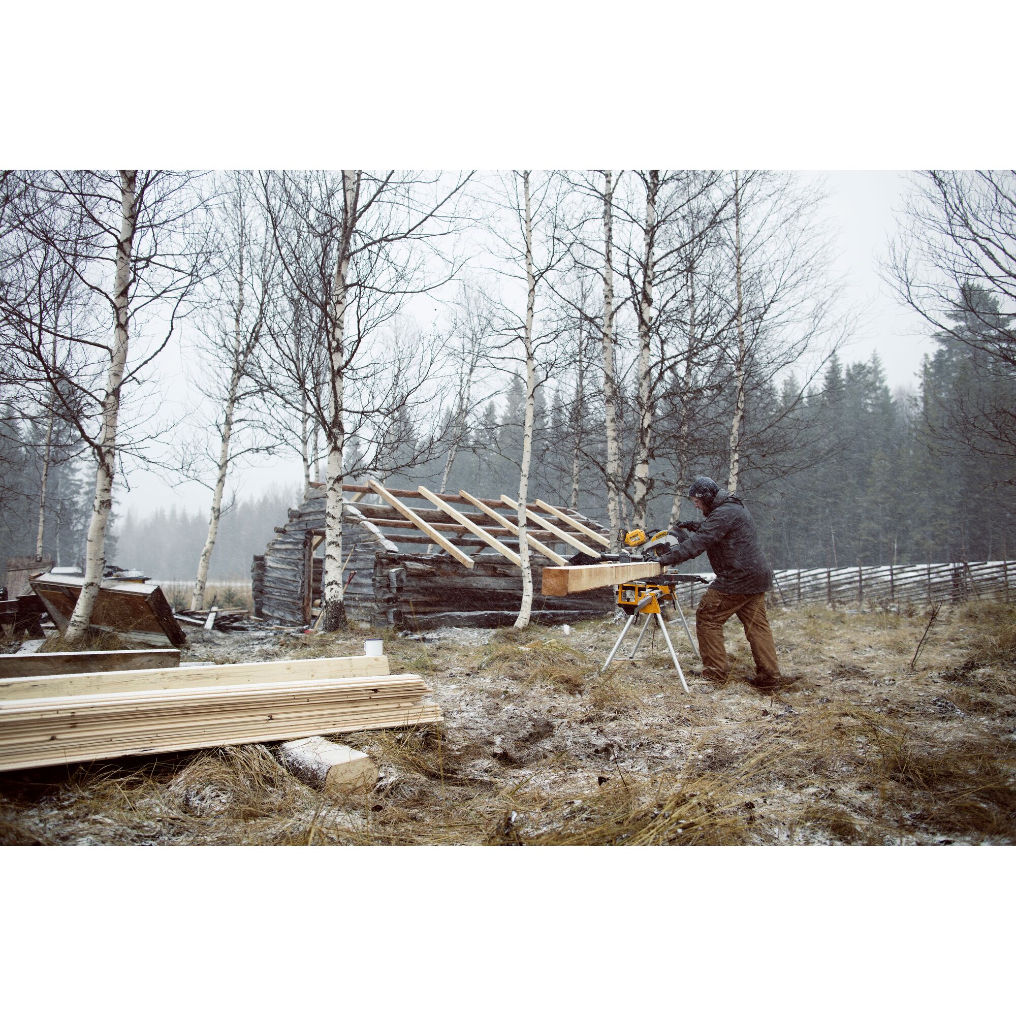 A person using a DEWALT miter saw on a stand outdoors in a snowy, rural area. There are stacks of wood and a partially constructed wooden structure surrounded by leafless trees.
