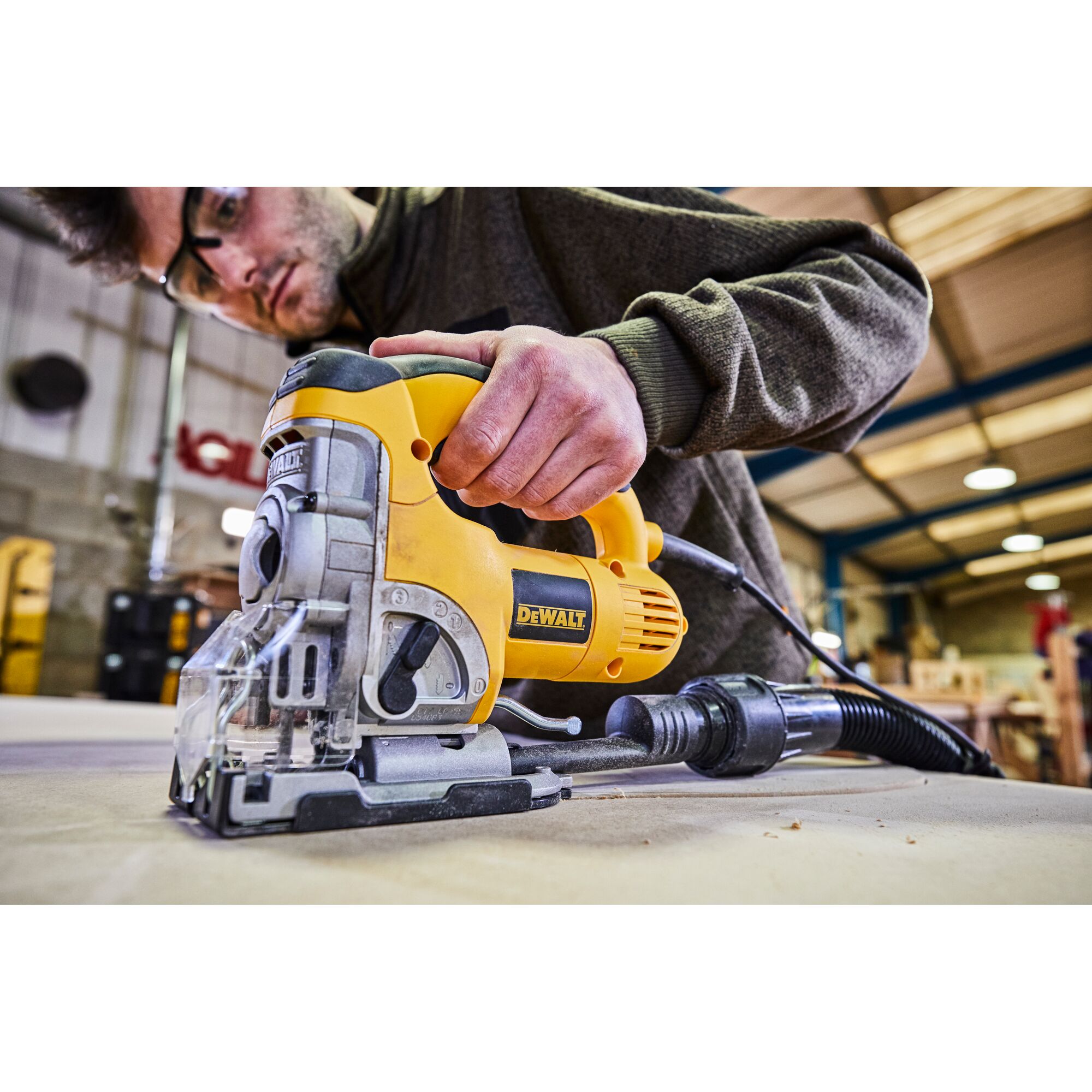 A close-up of a DEWALT DW331K jigsaw being operated on a piece of wood in a workshop. The person's hand is visible holding the tool, with dust extraction equipment attached. Workshop environment and wooden materials are in the background.