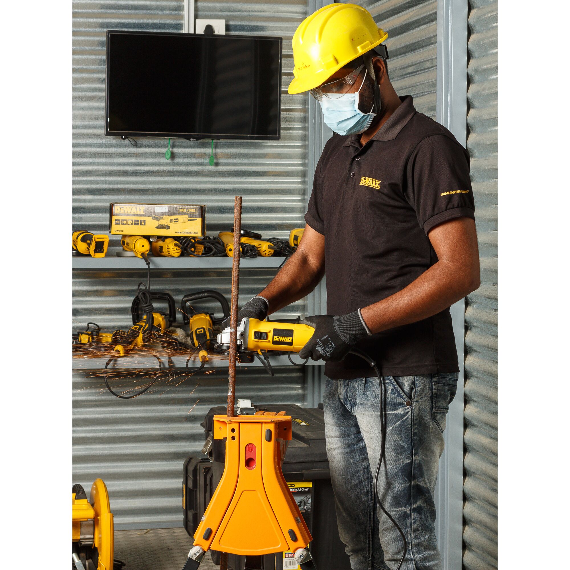 A person wearing a yellow hard hat and gloves is using a DEWALT DW802 power tool to cut a metal rod in an industrial workshop. The background shows a metal wall, shelves with various DEWALT tools, and a TV screen. The individual’s face is not visible.