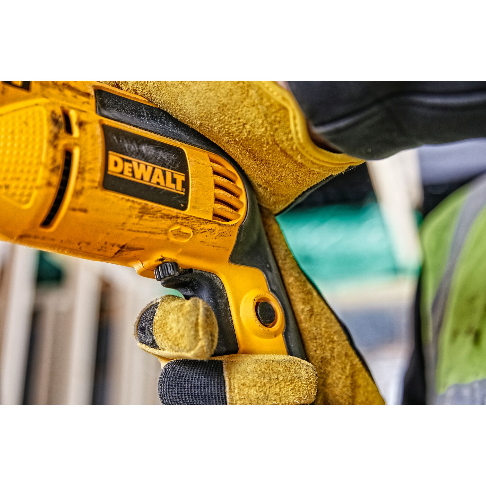 Close-up of a person wearing yellow work gloves holding a DEWALT electric drill at a construction site.