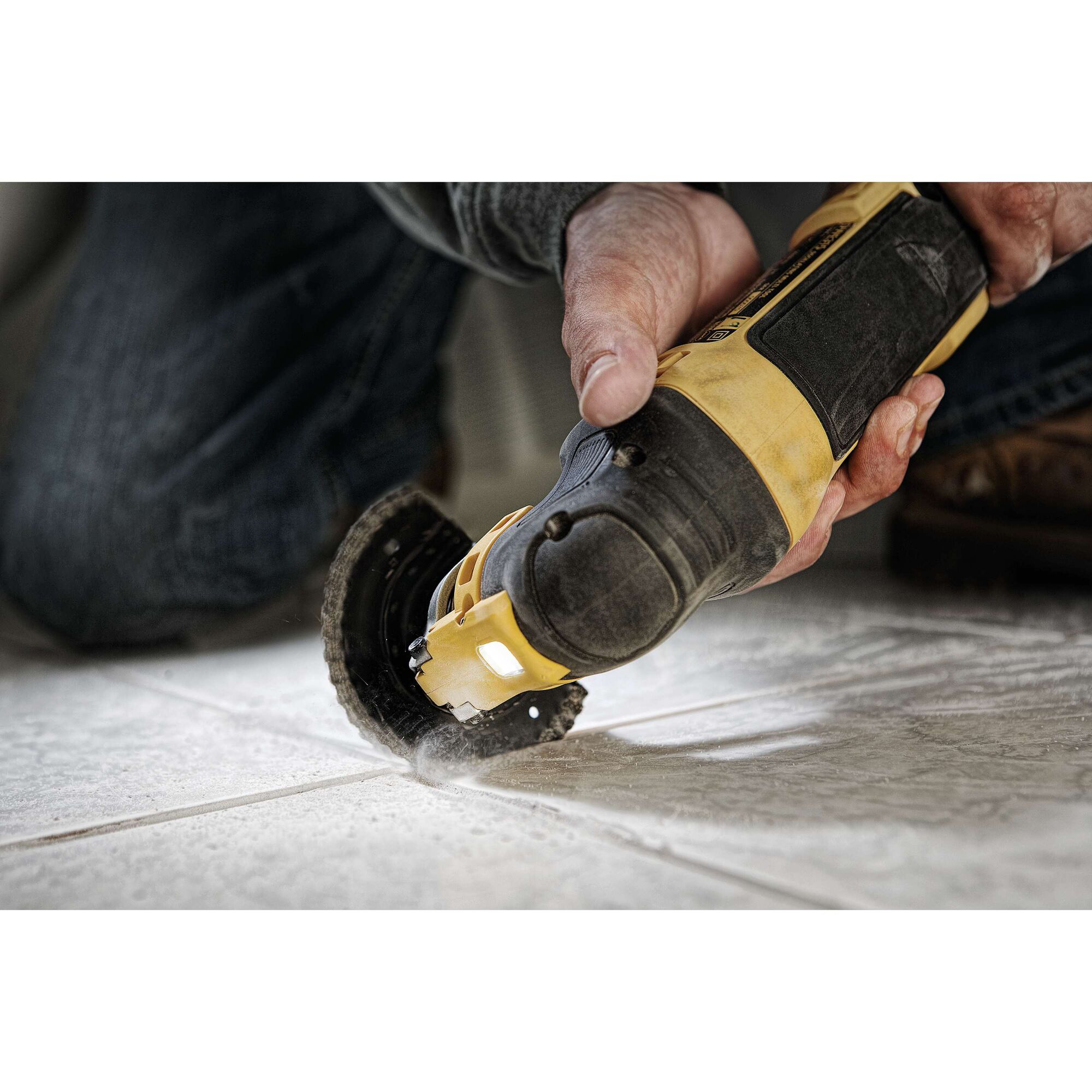 A person using a DEWALT DWE315 oscillating multi-tool to cut grout between tiles on a floor. The tool is yellow and black, and dust is visible as the cutting is performed.