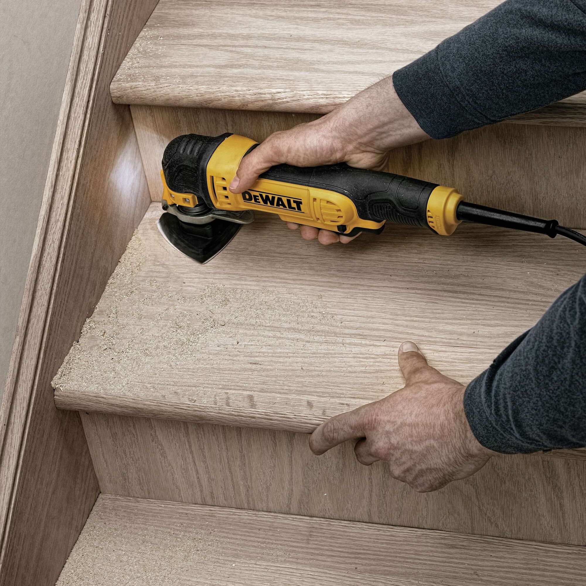 A person uses a DEWALT DWE315 oscillating multi-tool to sand the edge of a wooden stair step, producing sawdust.