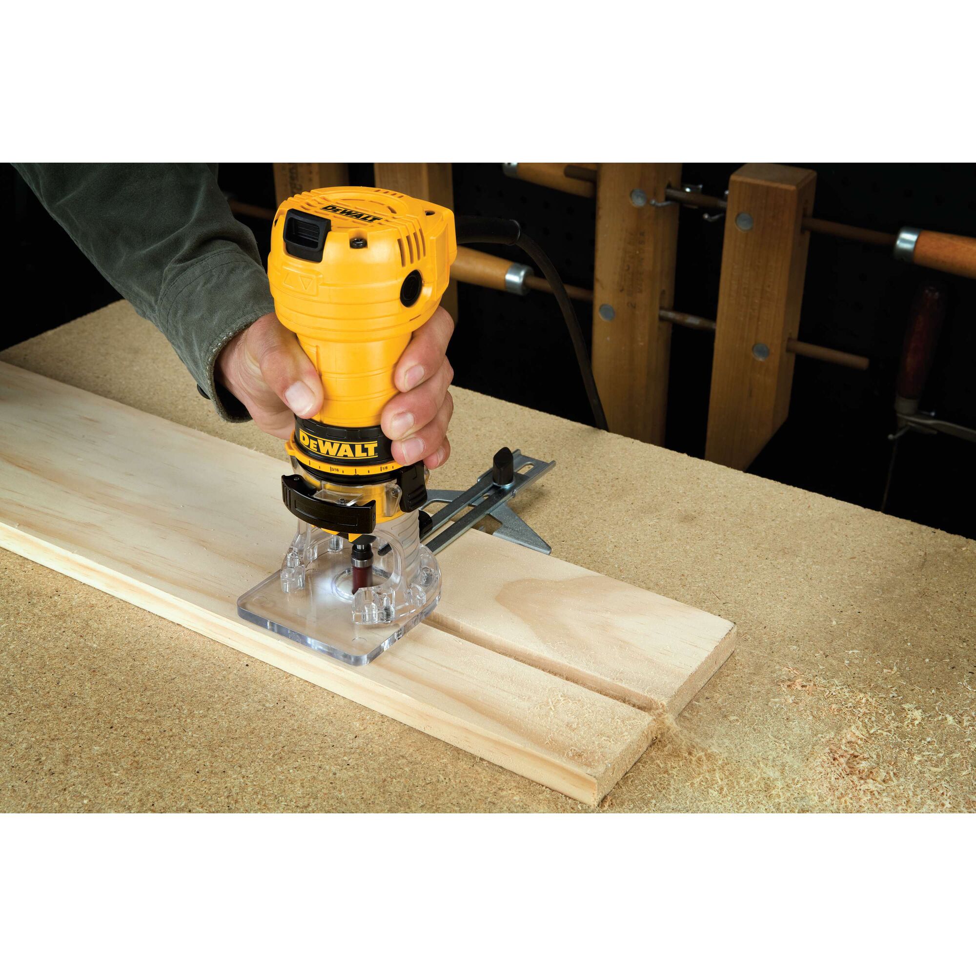 A person is using a DEWALT DWE6000 woodworking router to cut grooves into a piece of light-colored wood on a workbench.