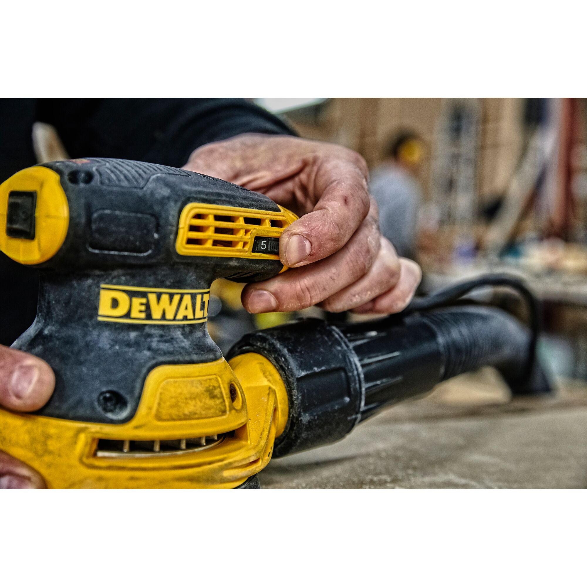 Close-up view of a person's hand adjusting a setting on a DEWALT power tool, specifically a yellow and black orbital sander, in a workshop environment.