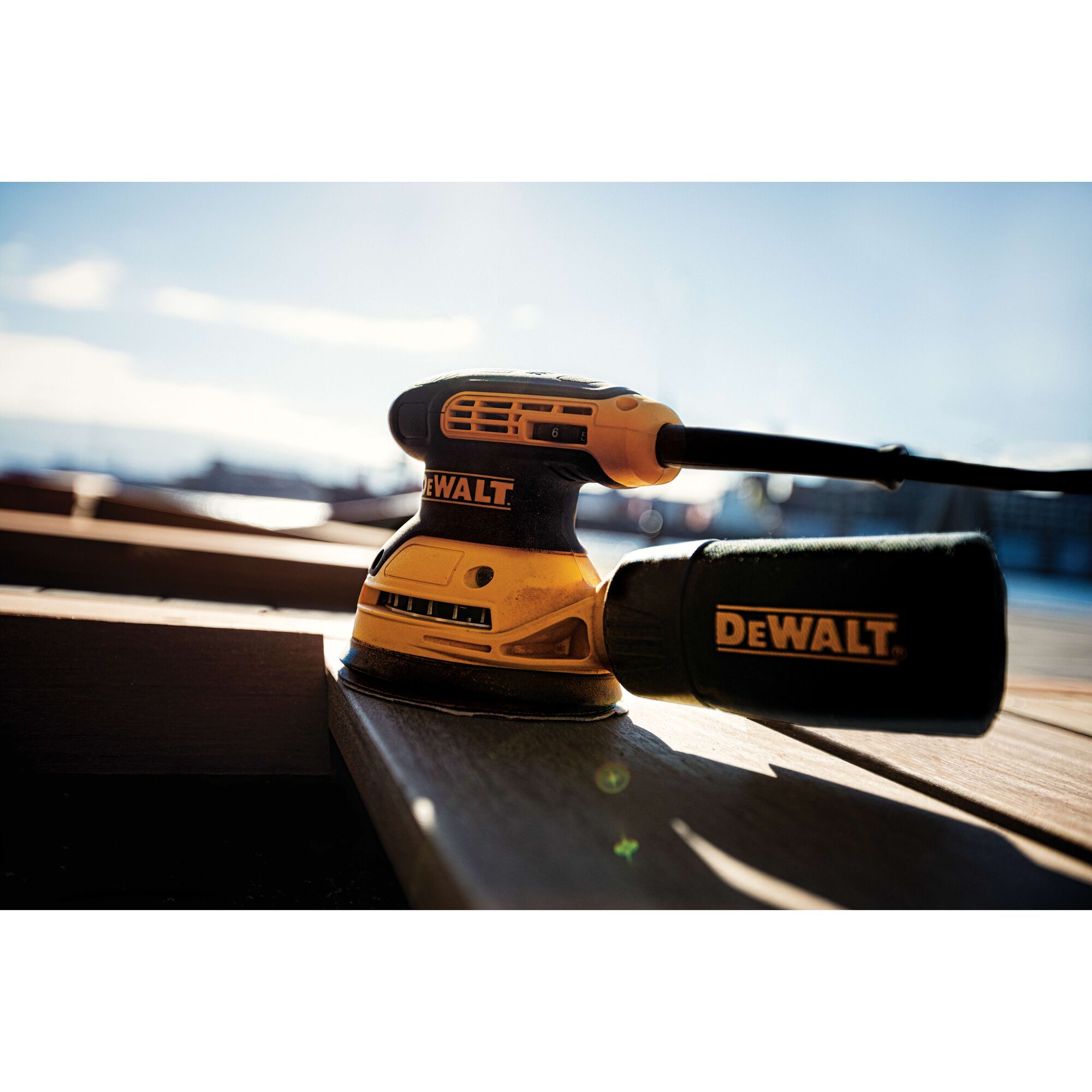 A DEWALT DWE6423 electric random orbital sander resting on a wooden surface outdoors, with sunlight illuminating the tool.