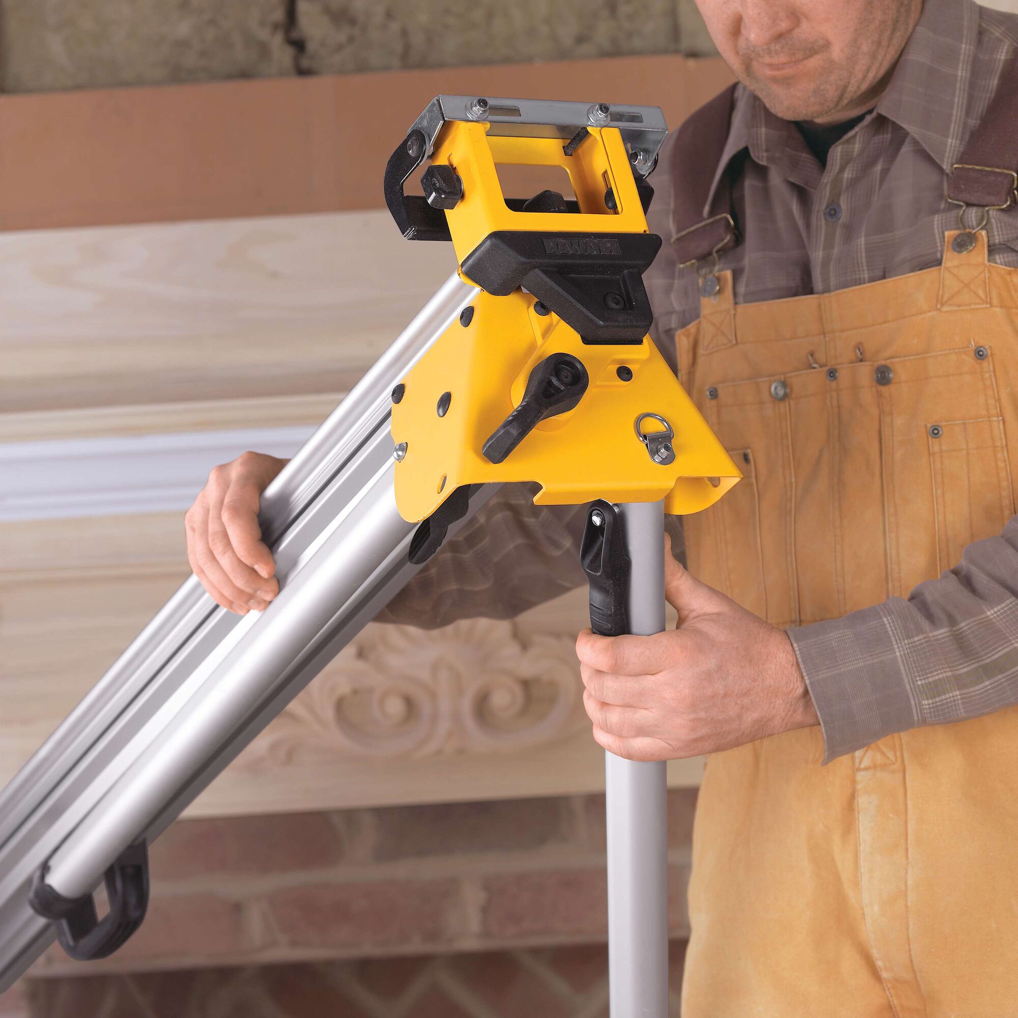 A person adjusting a DEWALT DWS780 miter saw stand with a yellow frame and metal legs, used for supporting woodworking tools. The person is wearing work clothes and is preparing the stand in front of a workshop setting.