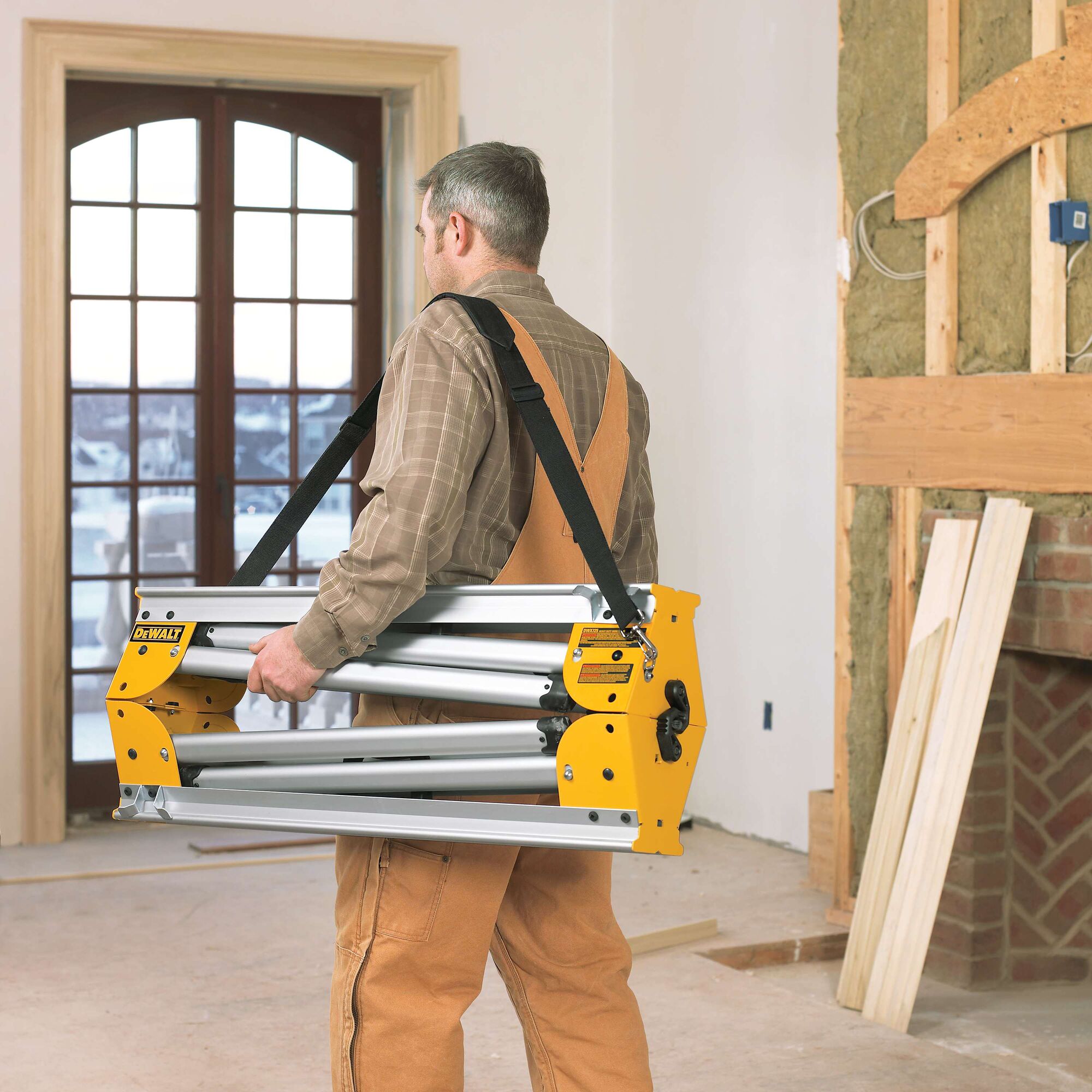 A person in work overalls carries a folded DEWALT miter saw stand with a shoulder strap inside a room under construction.