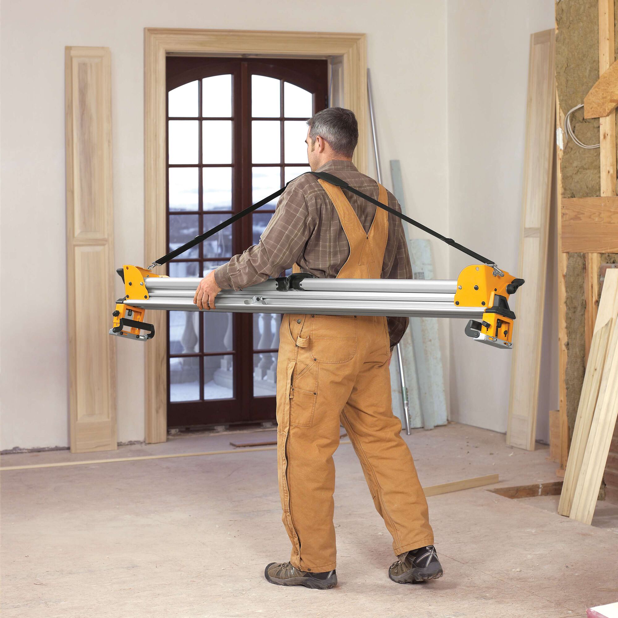 A person wearing brown overalls carries a DEWALT DWS780 photography equipment support rail with yellow ends and a strap in a room under construction.